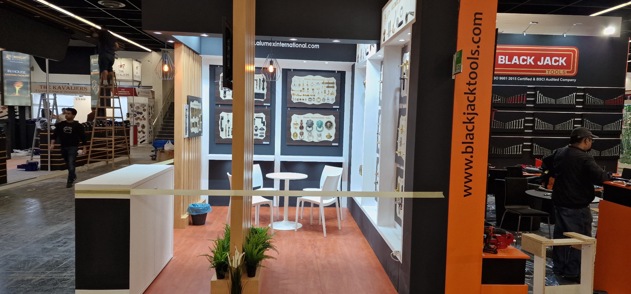 Stoisko targowe z ekspozycją okuć meblowych i drzwiowych, oświetlone lampami wiszącymi, z białymi krzesłami i stolikiem, oraz dekoracyjnymi roślinami w doniczkach.
