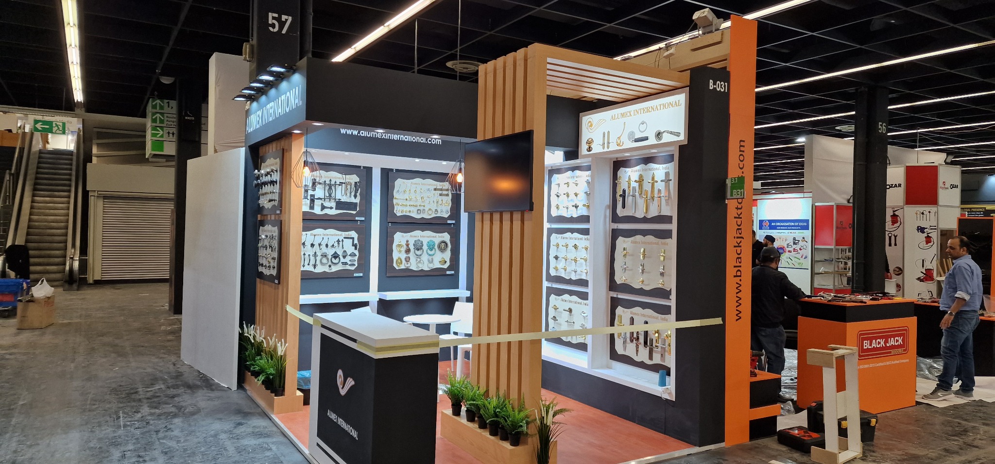 Ekspozycja stoiska targowego z elementami wyposażenia wnętrz, prezentująca kolekcję klamek i uchwytów do mebli w różnych stylach, z drewnianymi akcentami i roślinami doniczkowymi.