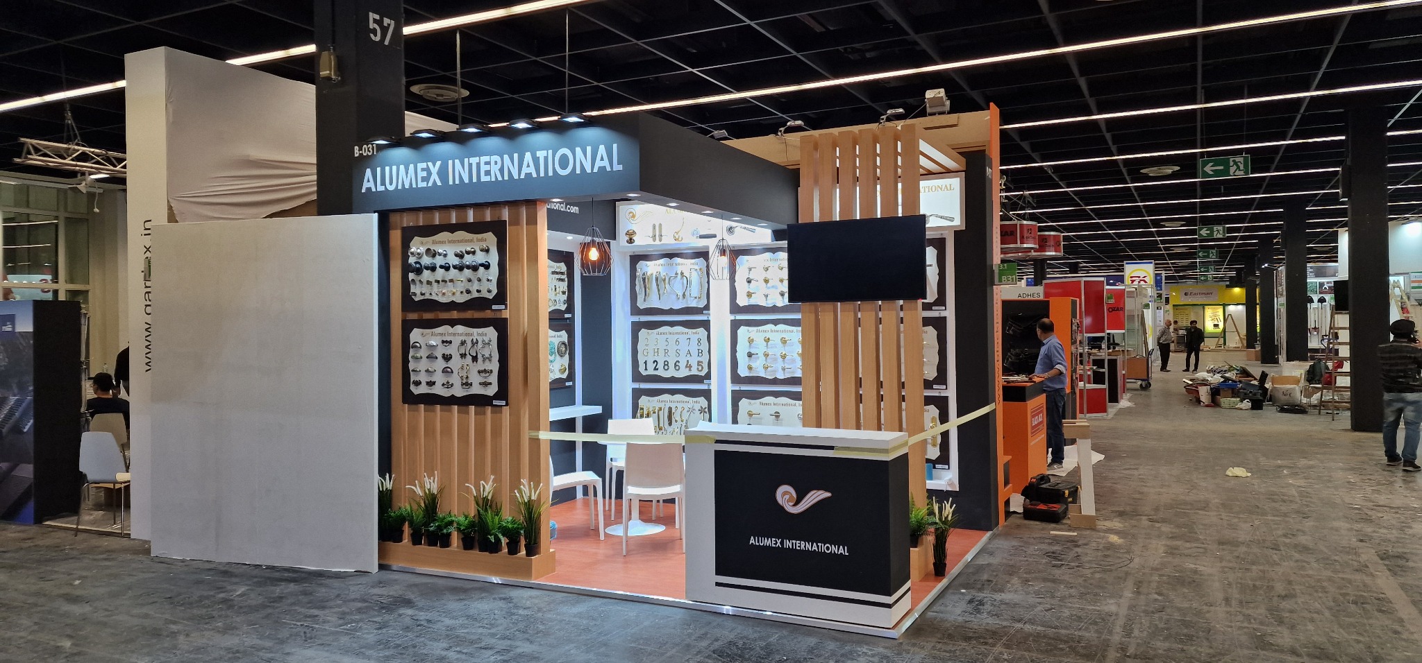 Stoisko targowe firmy Alumex International z ekspozycją okuć meblowych i drzwiowych, oświetlone lampami wiszącymi i z ekranem LCD, otoczone roślinami w doniczkach.