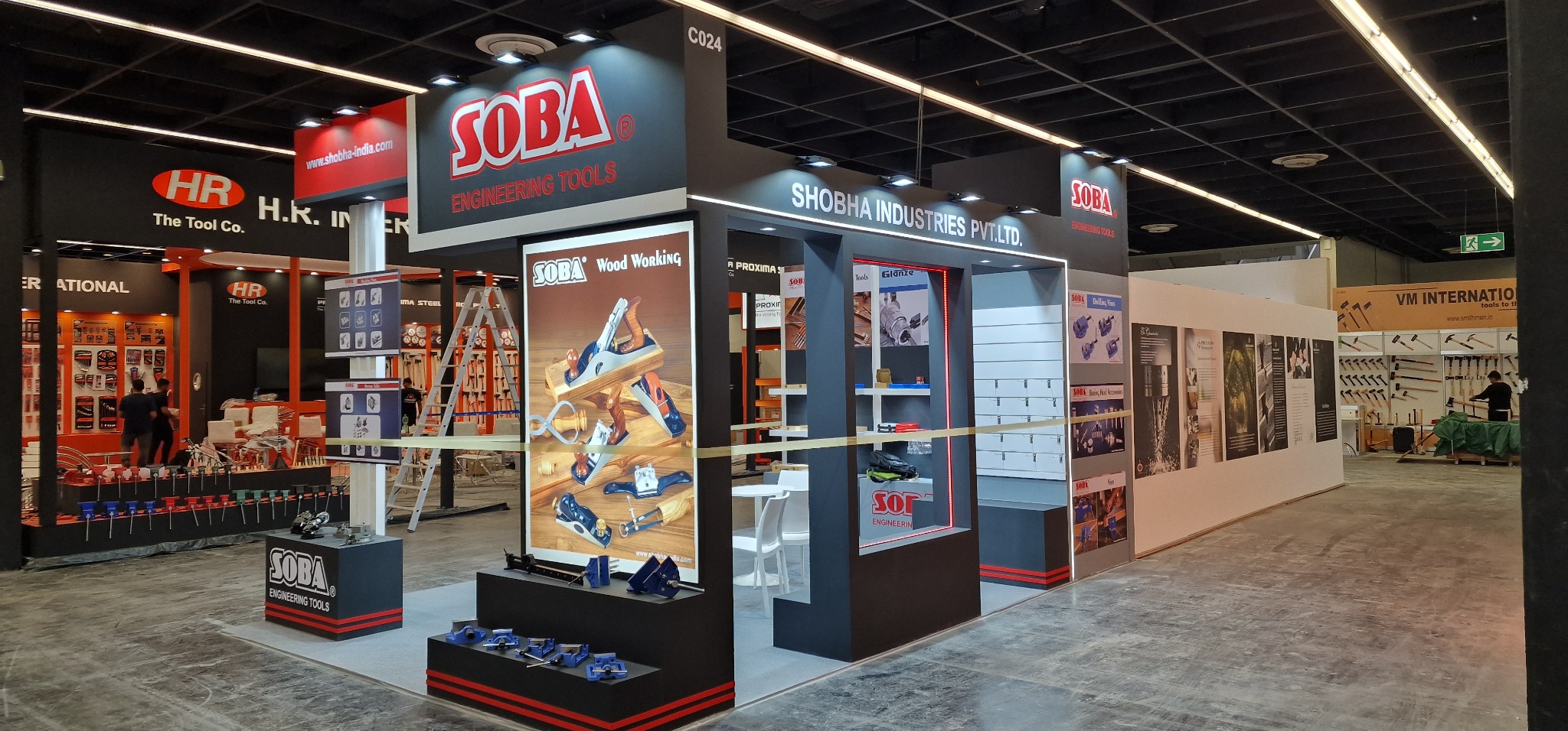 Widok stoiska targowego firmy SOBA prezentującego narzędzia do obróbki drewna, w tym strugi i ściski stolarskie, z ekspozycją na plakacie reklamowym i półkach.