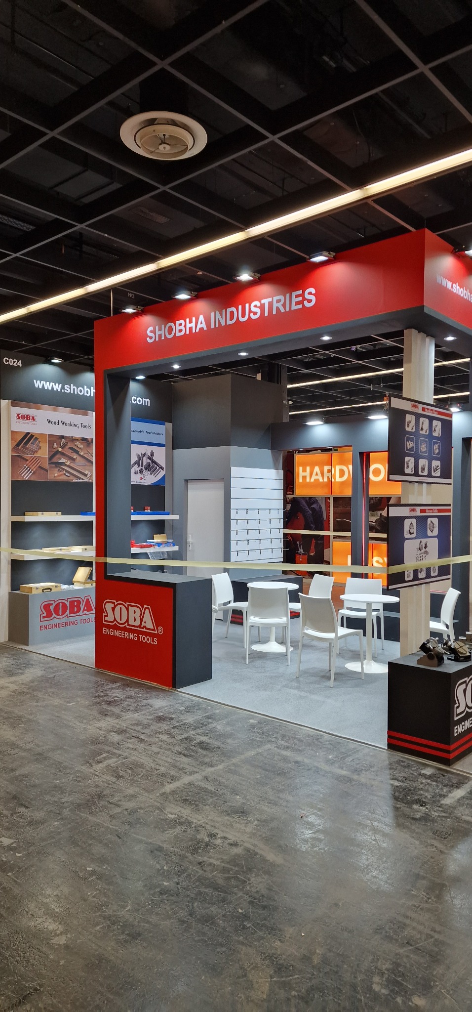 Stoisko targowe firmy Shobha Industries z ekspozycją narzędzi do obróbki drewna, prezentujące logo i ofertę, z białymi krzesłami i stolikami w przestrzeni ekspozycyjnej.