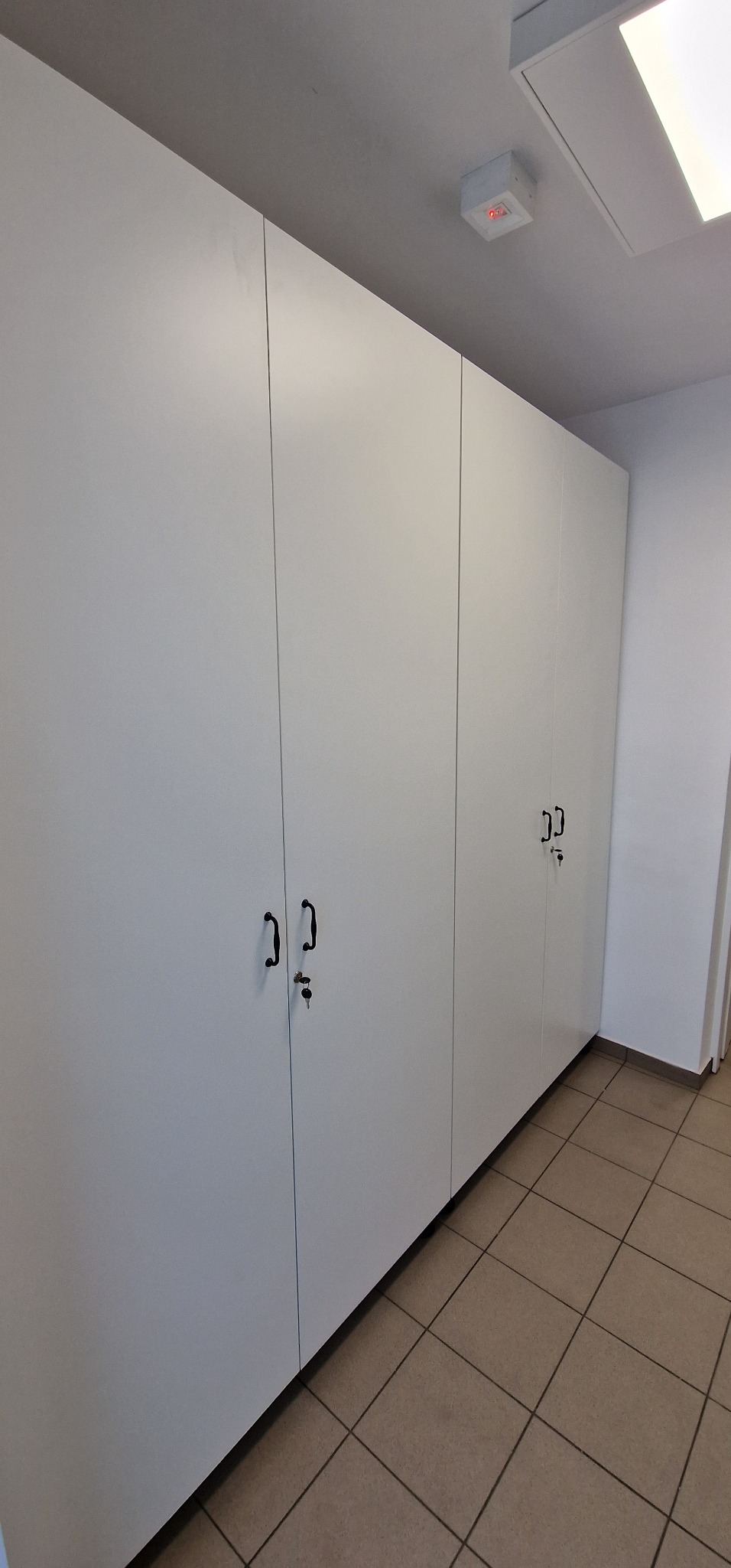 Długi rząd białych szaf z czarnymi uchwytami i kluczami w zamkach, ustawionych wzdłuż ściany z brązową podłogą z płytek.