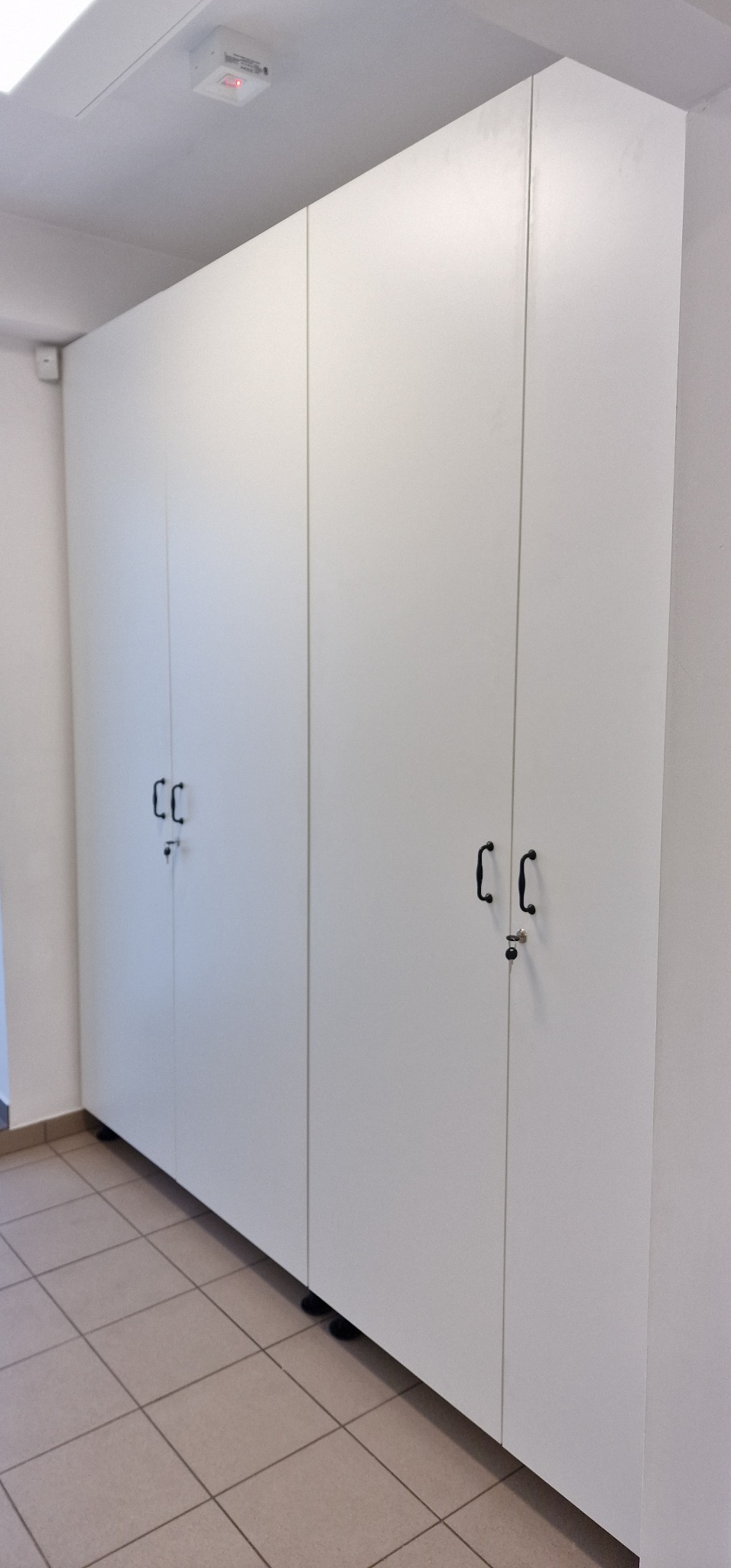 Biała, wysoka szafa z czterema drzwiami, z czarnymi uchwytami i zamkami, ustawiona wzdłuż ściany na kafelkowanej podłodze.