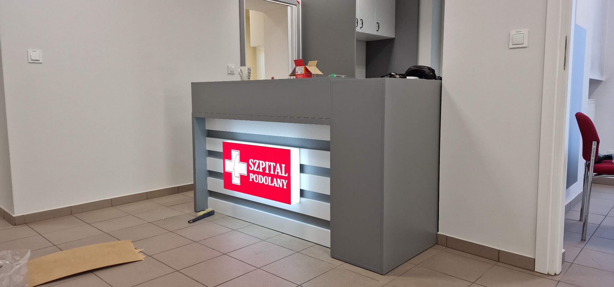 Szary, podświetlany panel recepcyjny z logo szpitala Podolany, widoczne elementy wykończenia wnętrza, w tle szafki wiszące.