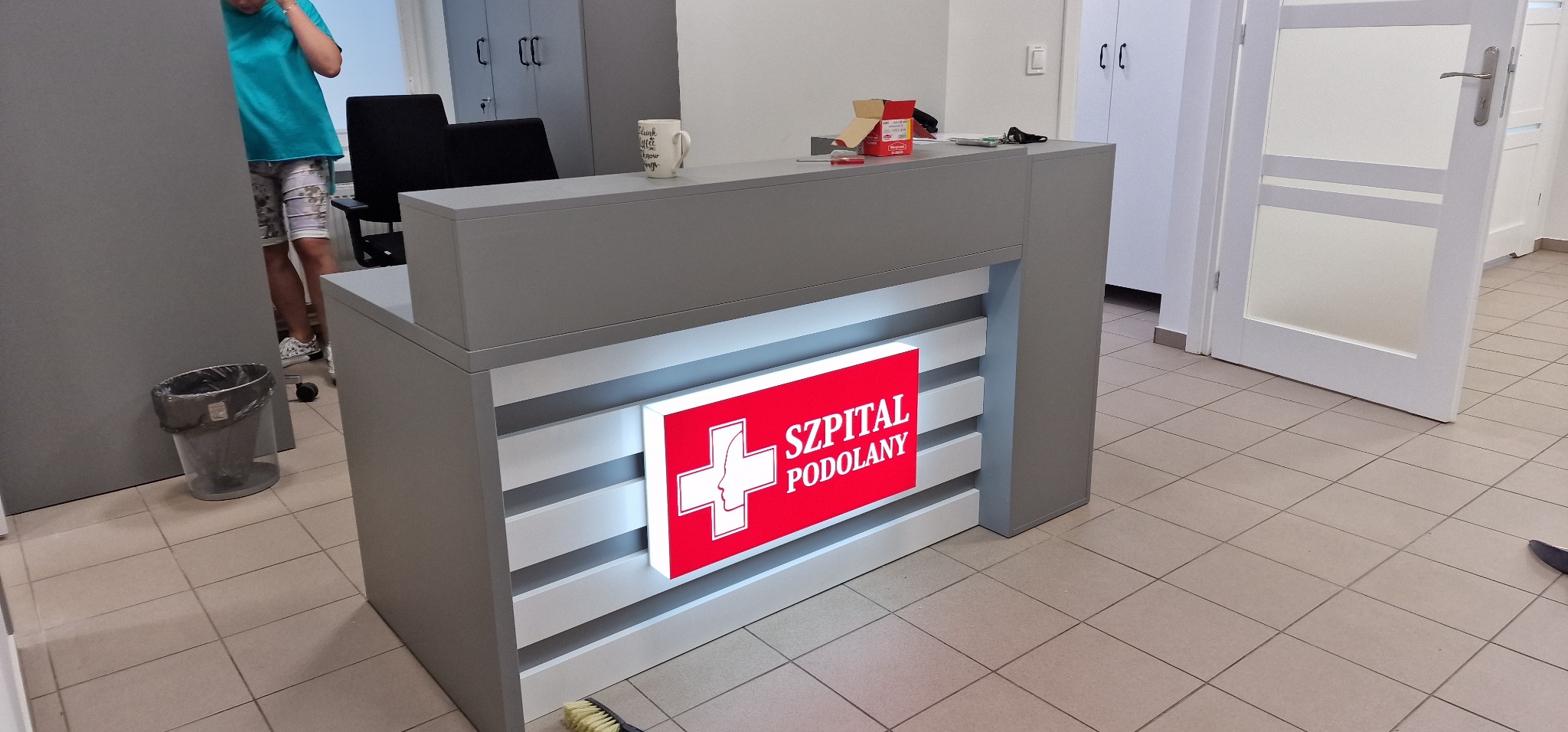 Szara lada recepcyjna z podświetlanym panelem 'Szpital Podolany', kubek i akcesoria na blacie, osoba w tle.