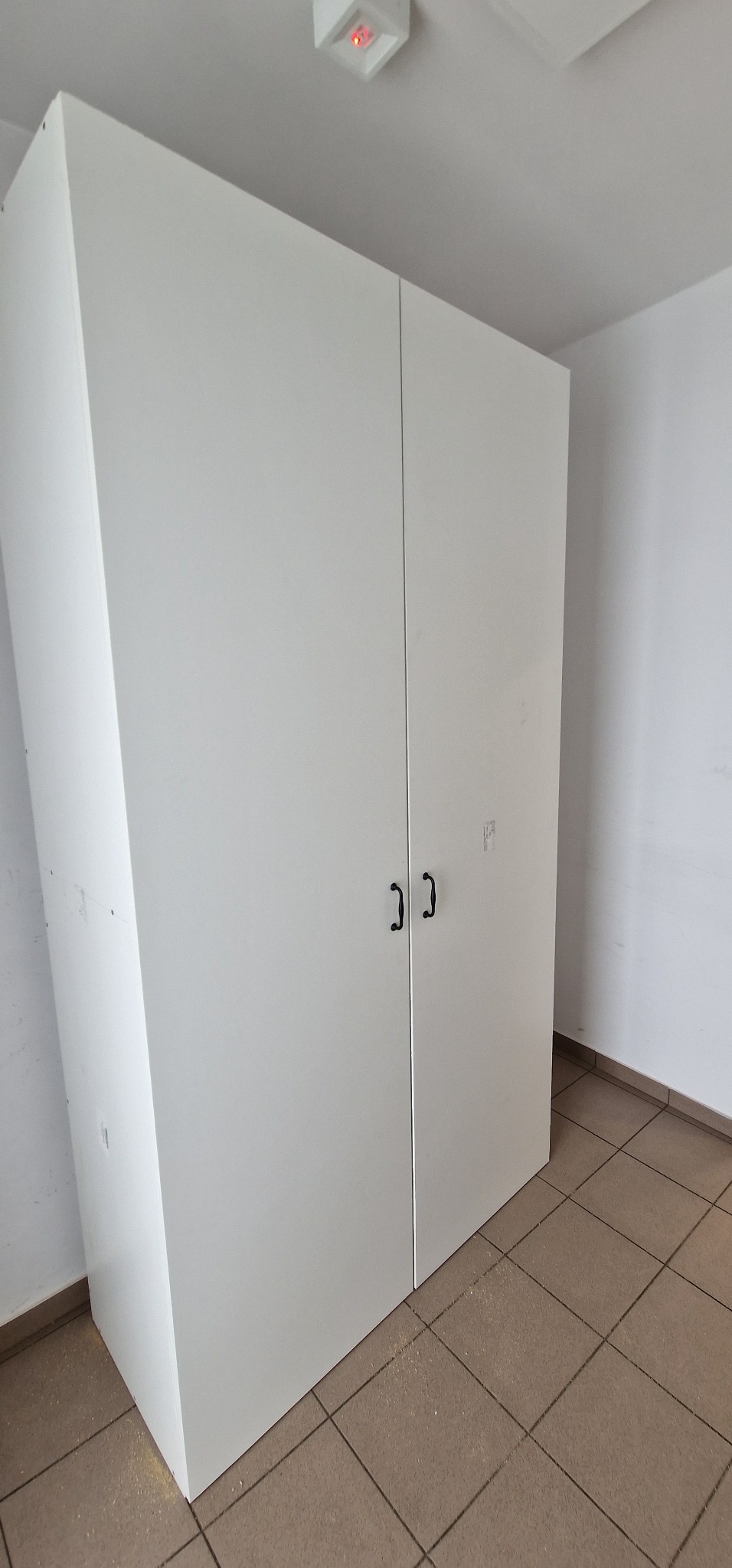 Biała, dwudrzwiowa szafa z ciemnymi uchwytami, widoczna z perspektywy podłogi z brązowych płytek, z widocznymi śladami po obróbce drewna.