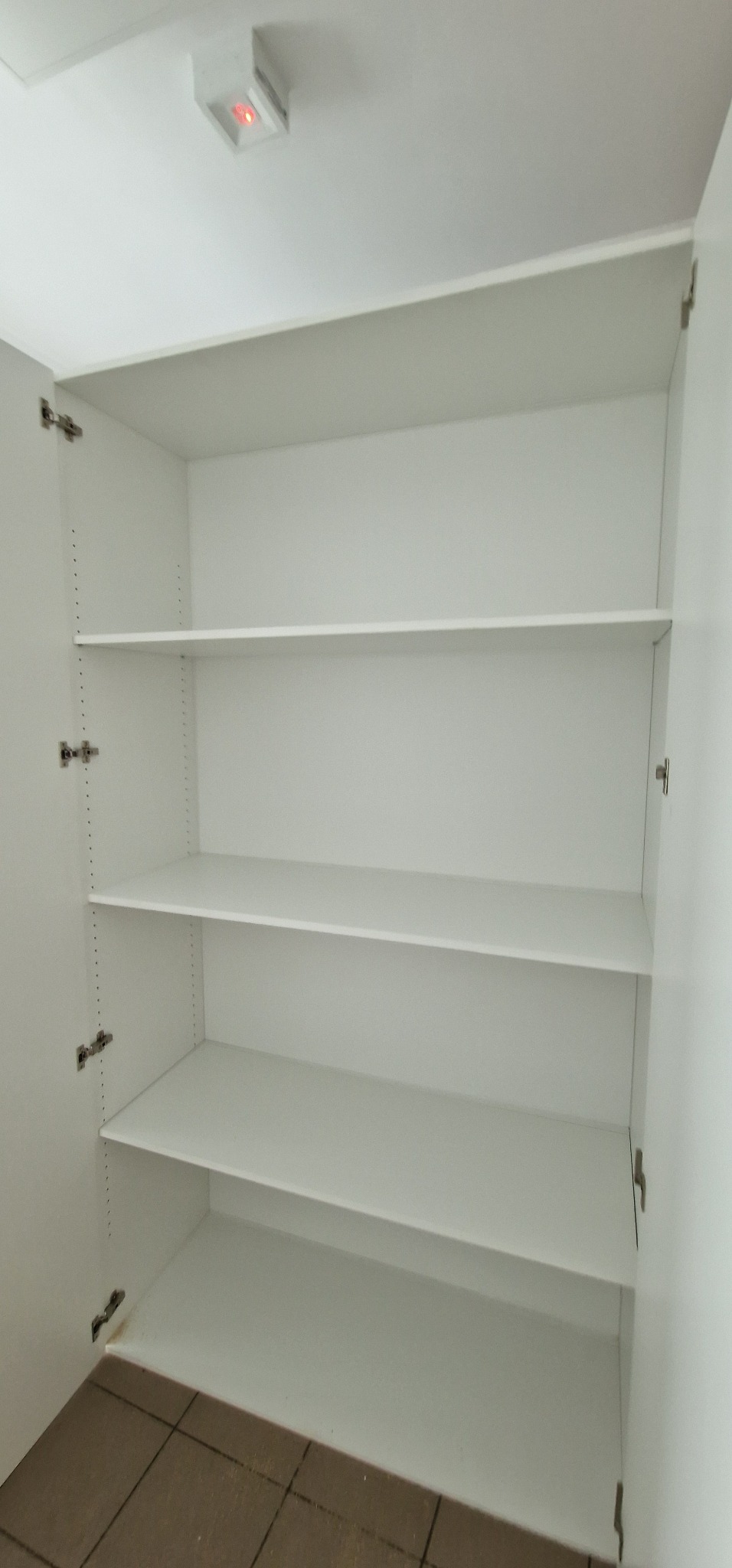 Wnętrze białej, pustej szafy wnękowej z regulowanymi półkami, widoczne otwory montażowe na bocznej ścianie, podłoga z brązowych płytek w dolnej części kadru.