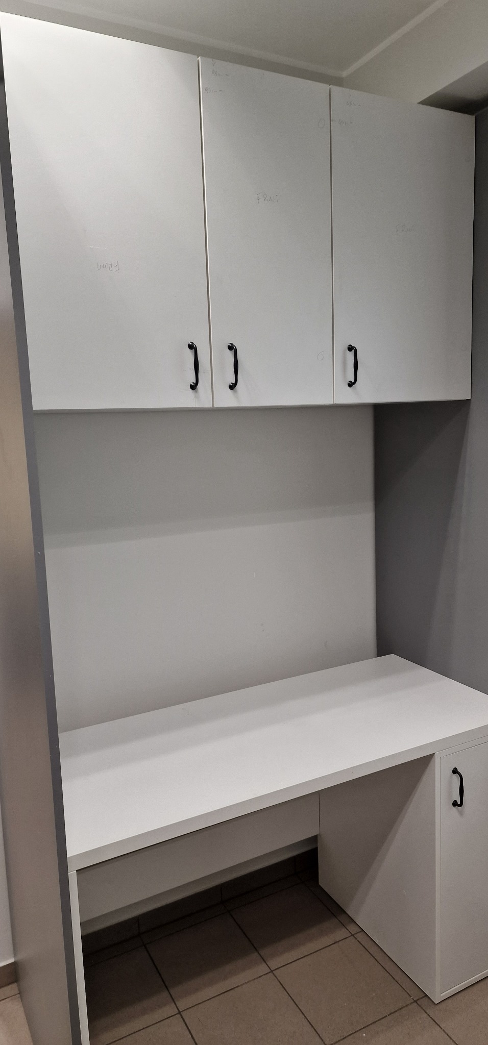 Białe biurko z szafkami wiszącymi, czarne uchwyty, widoczne notatki ołówkiem na frontach, niedokończony montaż.
