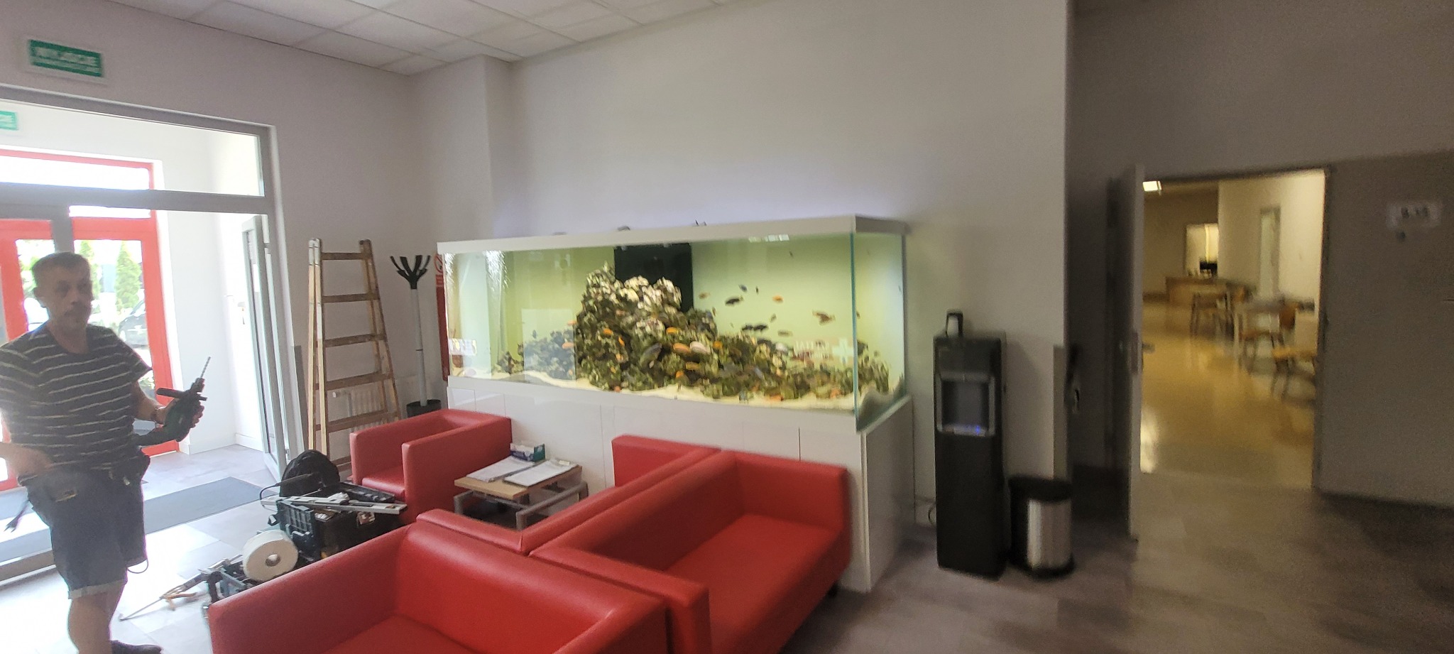 Mężczyzna z wiertarką w ręku stoi w pomieszczeniu z akwarium, czerwonymi sofami i drabiną, prawdopodobnie podczas montażu lub naprawy