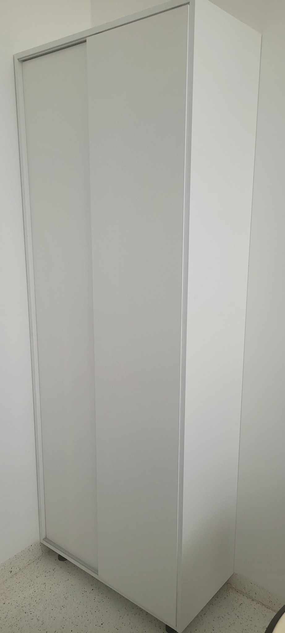 Biała szafa z przesuwnymi drzwiami, widoczna z boku, na tle białej ściany i podłogi z lastryko.