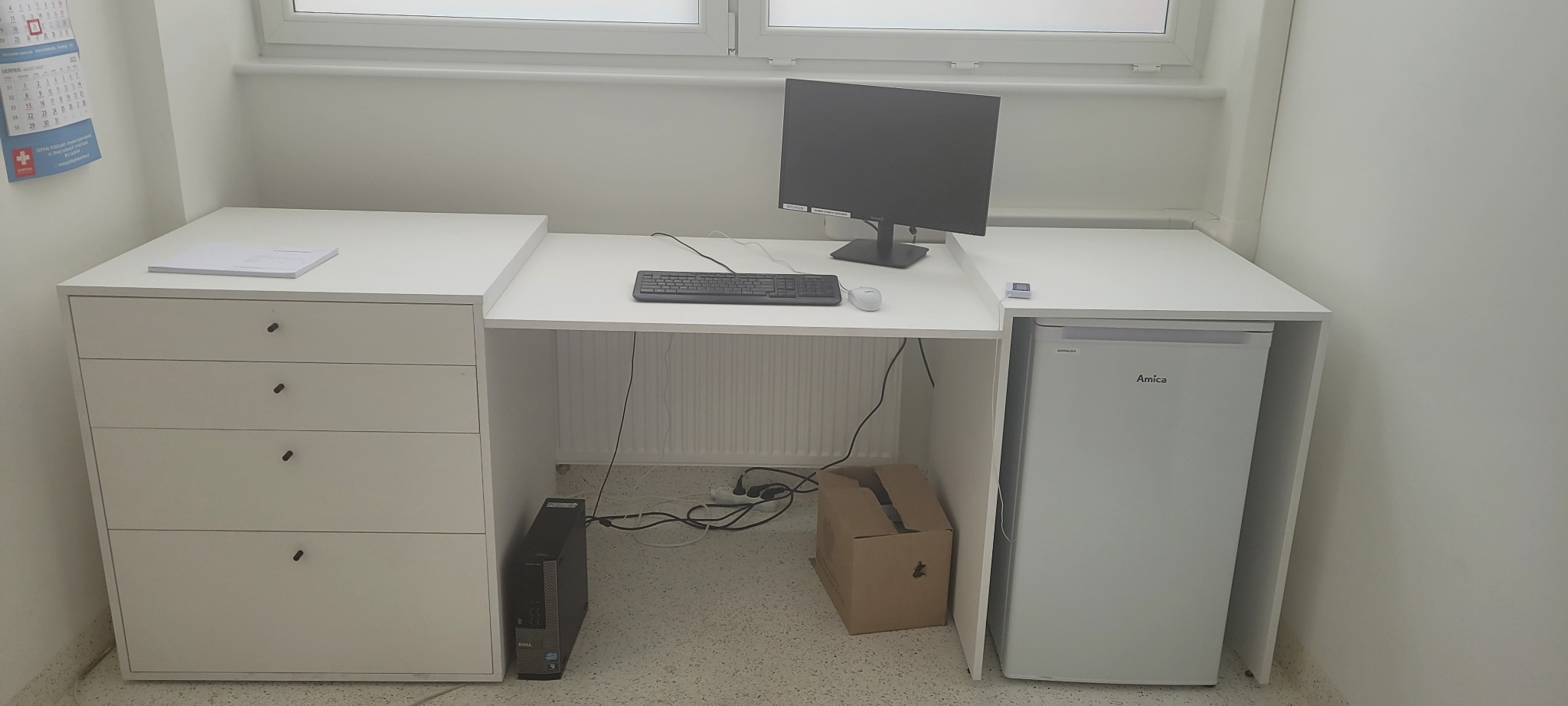 Białe biurko z szufladami i wbudowaną lodówką Amica, komputer stacjonarny i monitor na blacie, widoczny kabel zasilający i karton na podłodze