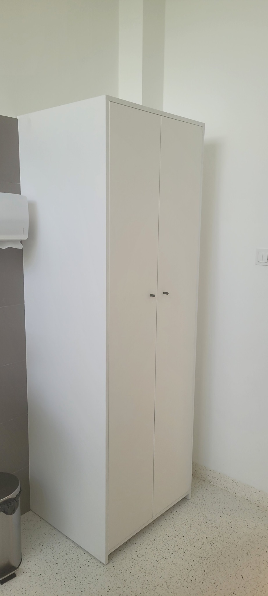 Biała, dwudrzwiowa szafa z minimalistycznymi uchwytami na tle jasnej ściany i podłogi z lastryko, obok szarej ściany z dozownikiem papieru i koszem na śmieci.