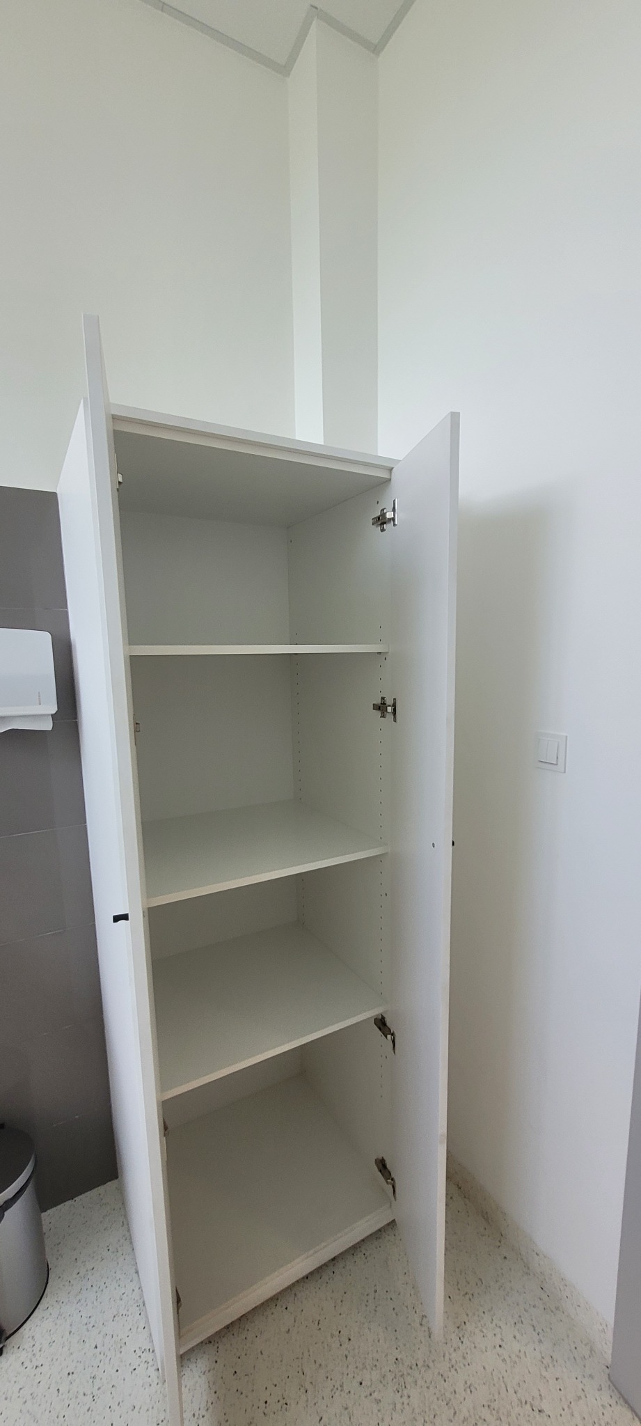 Wysoka, biała szafka z otwartymi drzwiami, widoczne cztery półki wewnątrz, zamontowana w pomieszczeniu z szarą ścianą i jasną podłogą.
