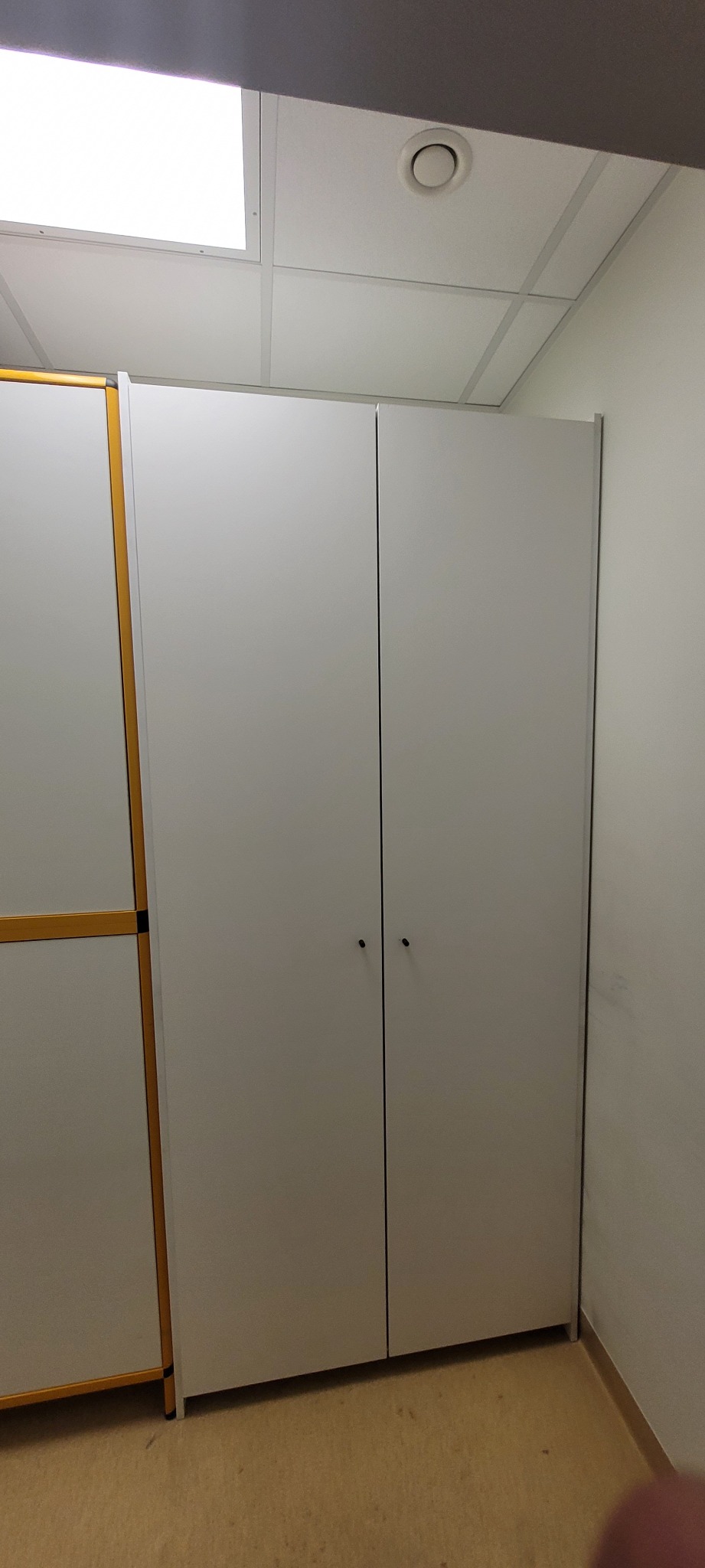 Zamknięta, dwudrzwiowa szafa z jasnymi frontami i małymi, okrągłymi uchwytami na tle biurowego wnętrza z podwieszanym sufitem.