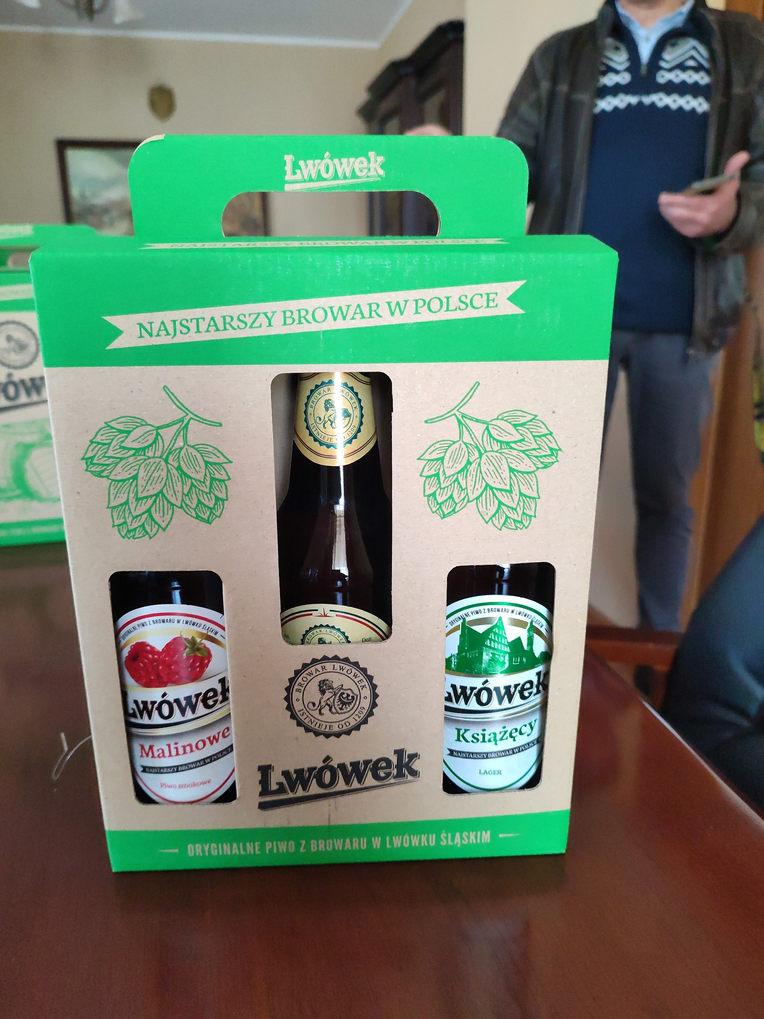 Zestaw trzech butelek piwa z browaru Lwówek w ozdobnym, zielonym kartonie z uchwytem, prezentujący smaki: malinowe, Książęcy Lager i klasyczne. Widoczny fragment osoby w tle.