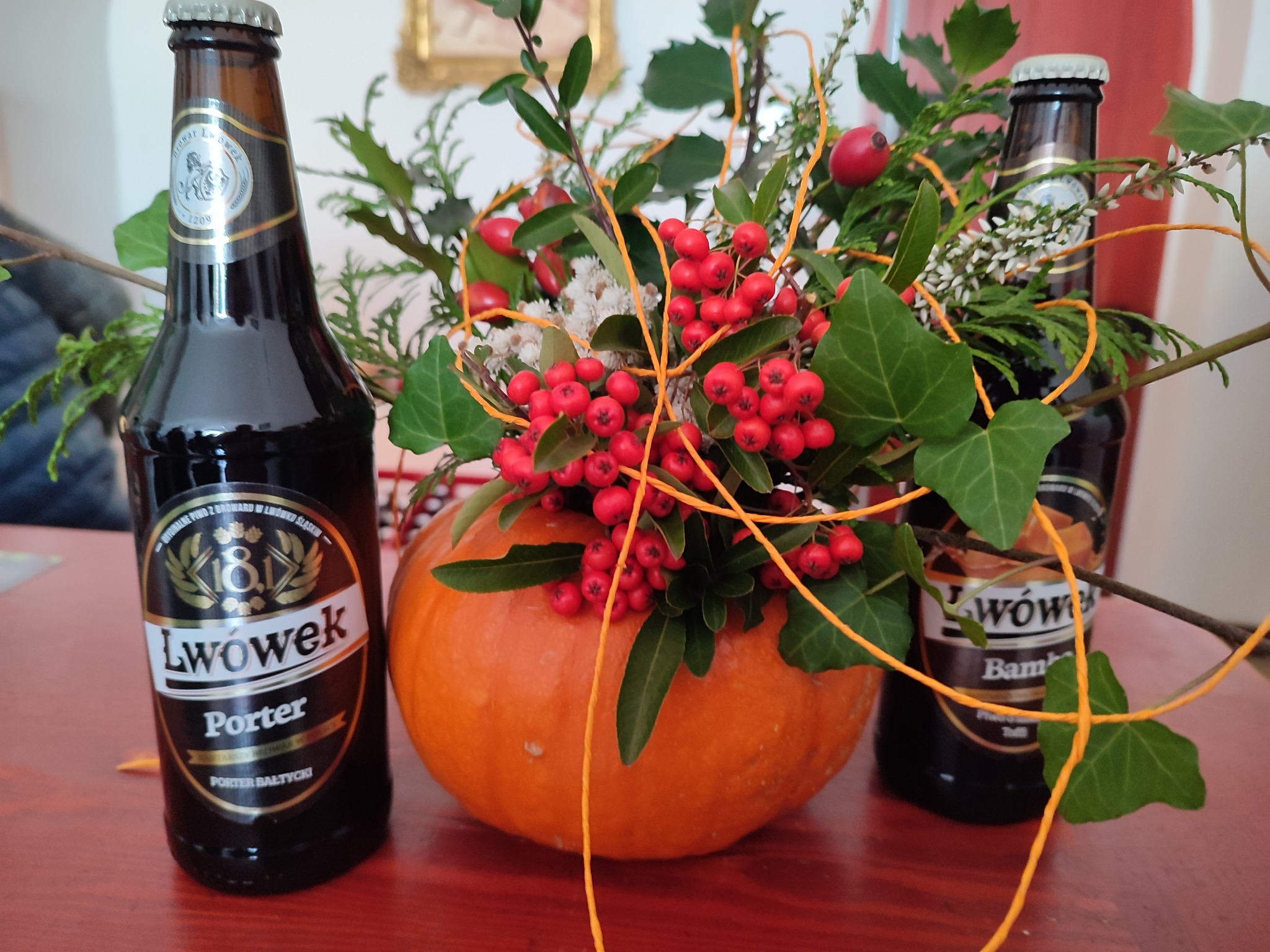 Dekoracja stołu z dyni, gałązek, czerwonych owoców i dwóch butelek ciemnego piwa Lwówek Porter i Lwówek Bamberg, tworząca jesienną kompozycję.