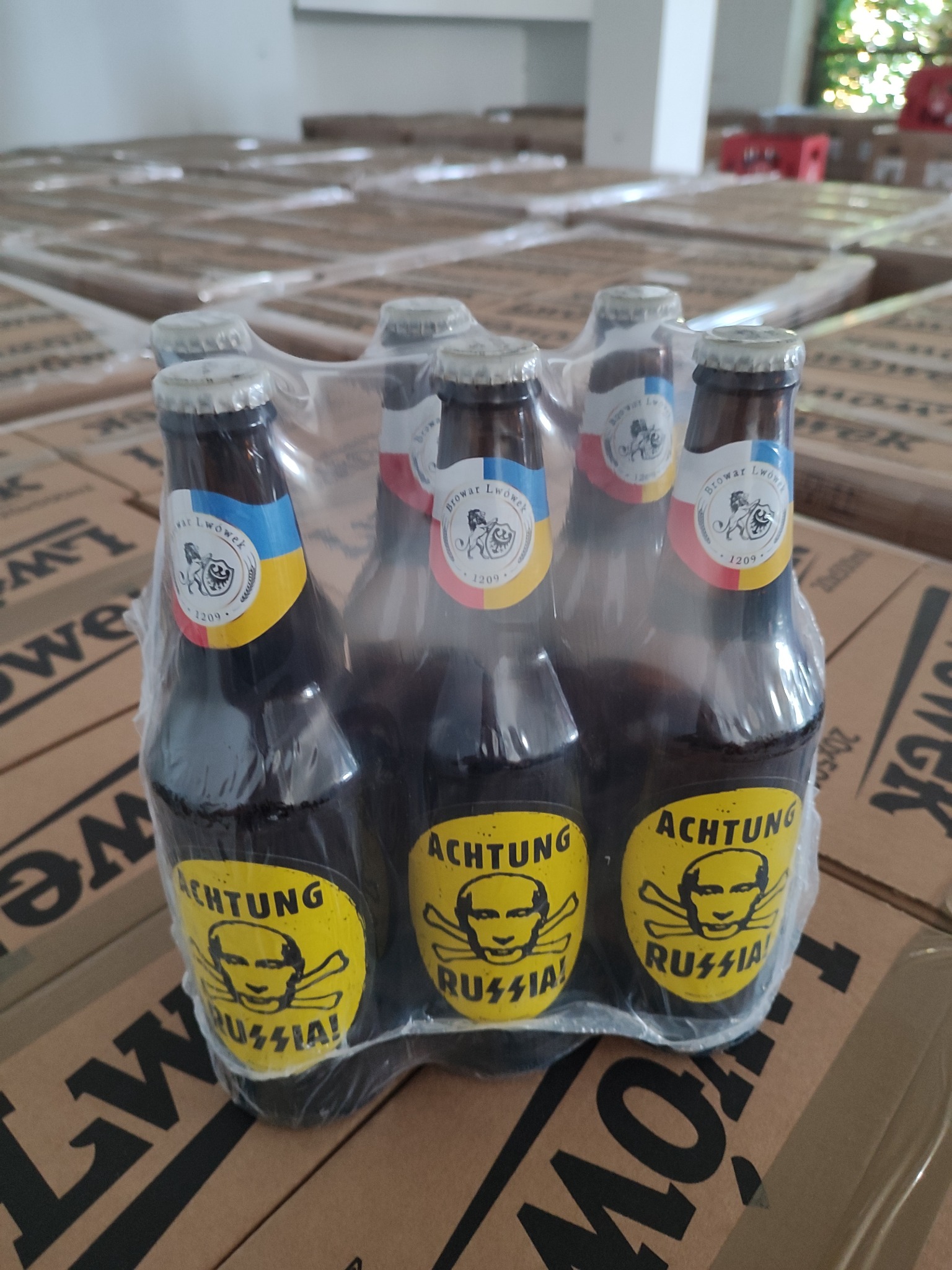Sześciopak piwa Browar Lwówek w folii, z naklejkami 'Achtung Russia!' z wizerunkiem czaszki Putina, ustawiony na kartonach z napisem 'Lwówek', w tle magazyn z paletami piwa.