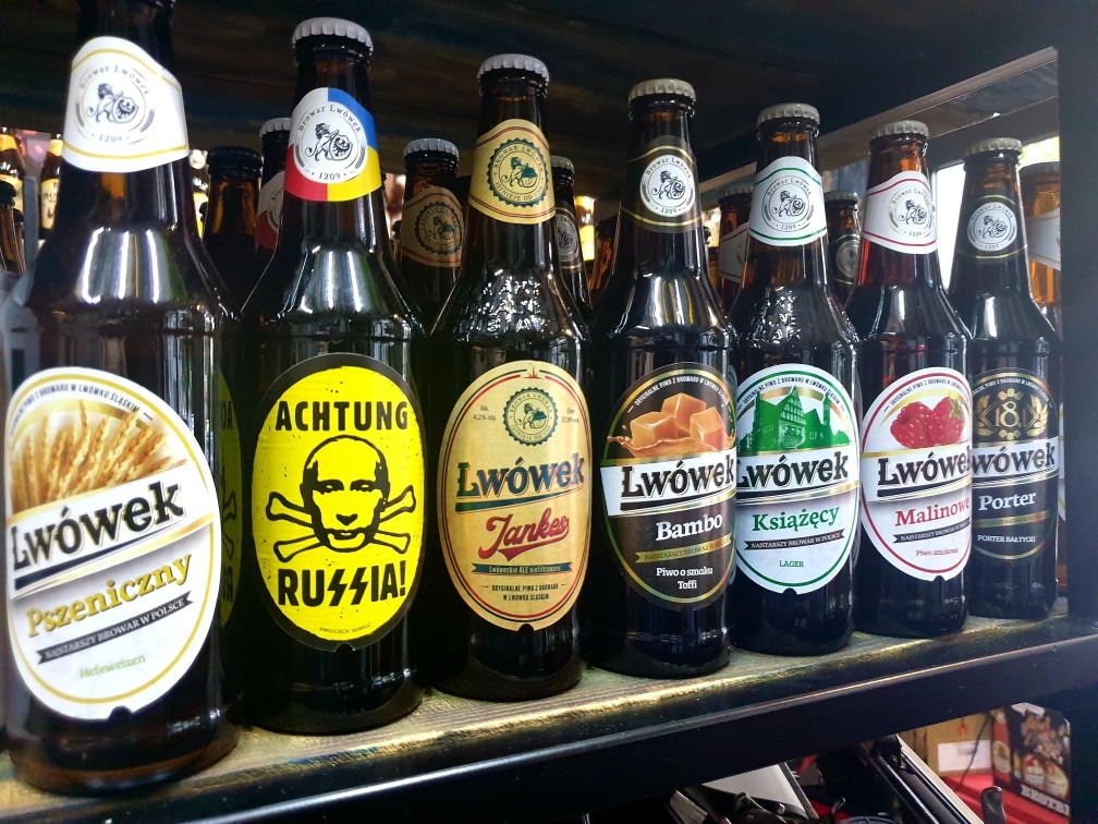 Różne rodzaje piwa Lwówek na półce, w tym pszeniczne, z malinami, porter i piwo o smaku toffi, jedno z piw z kontrowersyjną etykietą 'Achtung Russia' z wizerunkiem czaszki.