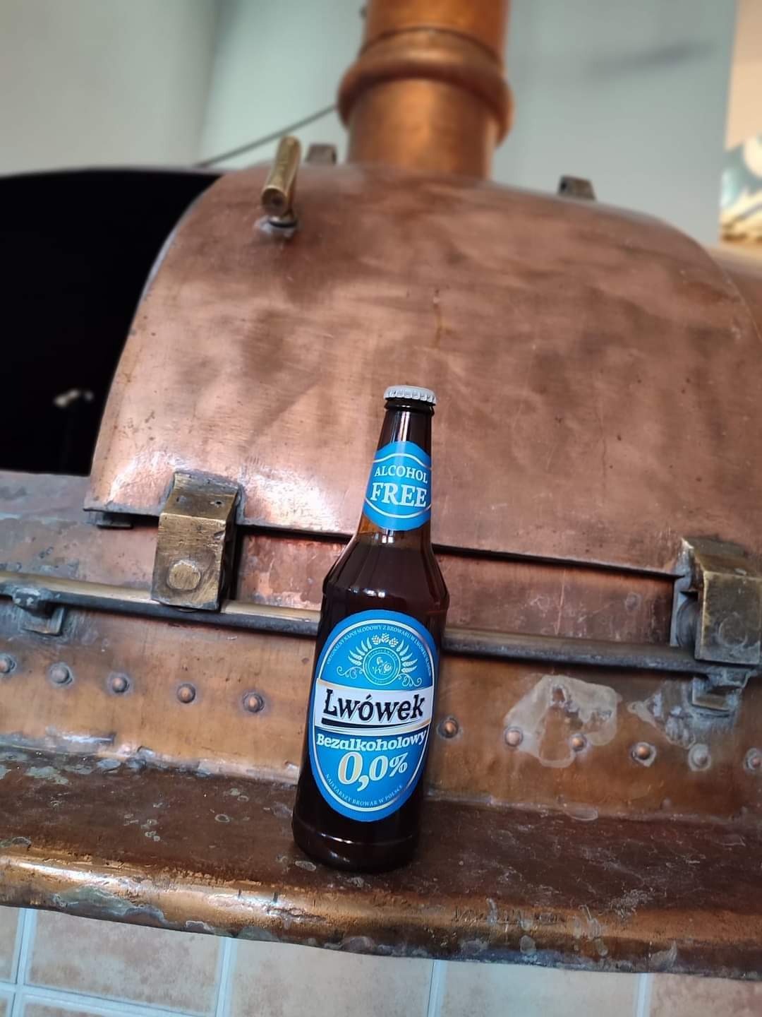 Butelka bezalkoholowego piwa Lwówek 0,0% na tle miedzianego kotła warzelnego z nitami i mosiężnymi zaworami.