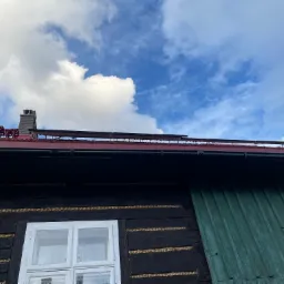 Panele fotowoltaiczne na dachu zabytkowego, drewnianego domu z oknami i kominem na tle błękitnego nieba z chmurami. Widok z dołu.