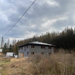 Sunrise-Technology JAKUB WITOS - Szary dom z panelami słonecznymi na dachu w trakcie montażu, widoczny zarośnięty teren i samochód dostawczy w tle, pochmurne niebo.