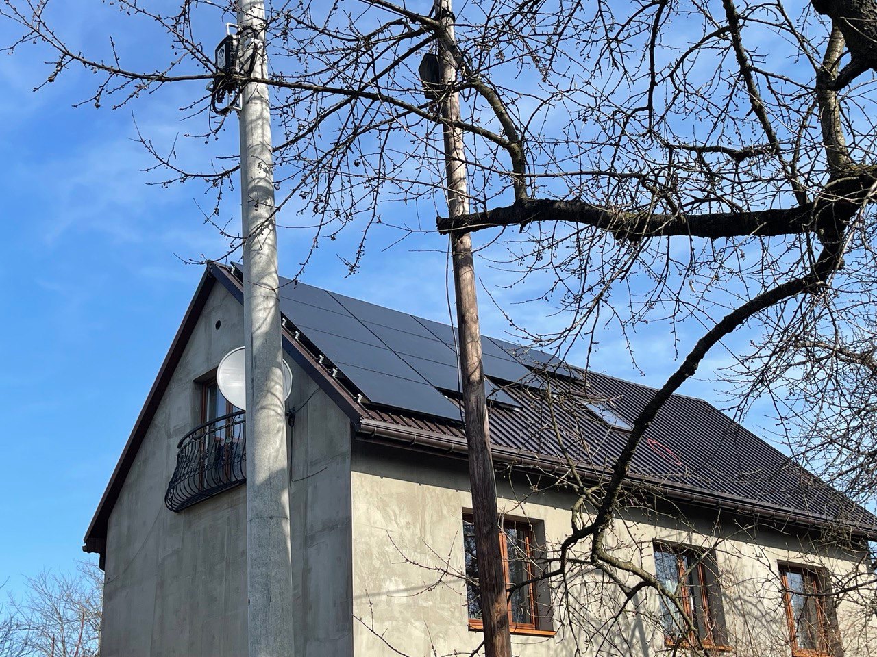 Dom jednorodzinny z panelami słonecznymi na dachu, widoczne z poziomu ulicy, z przewodami elektrycznymi i drzewami bez liści na pierwszym planie.