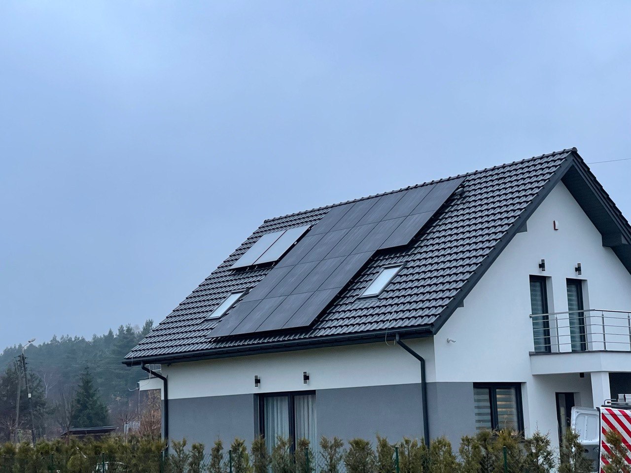 Instalacja paneli słonecznych na dachu dwukondygnacyjnego domu z szarej dachówki, widok z dołu w pochmurny dzień.