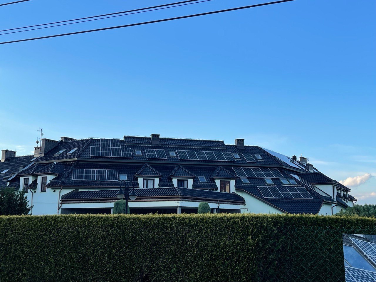 Duży dom z ciemnym dachem, na którym zamontowano panele słoneczne; widok zza żywopłotu.