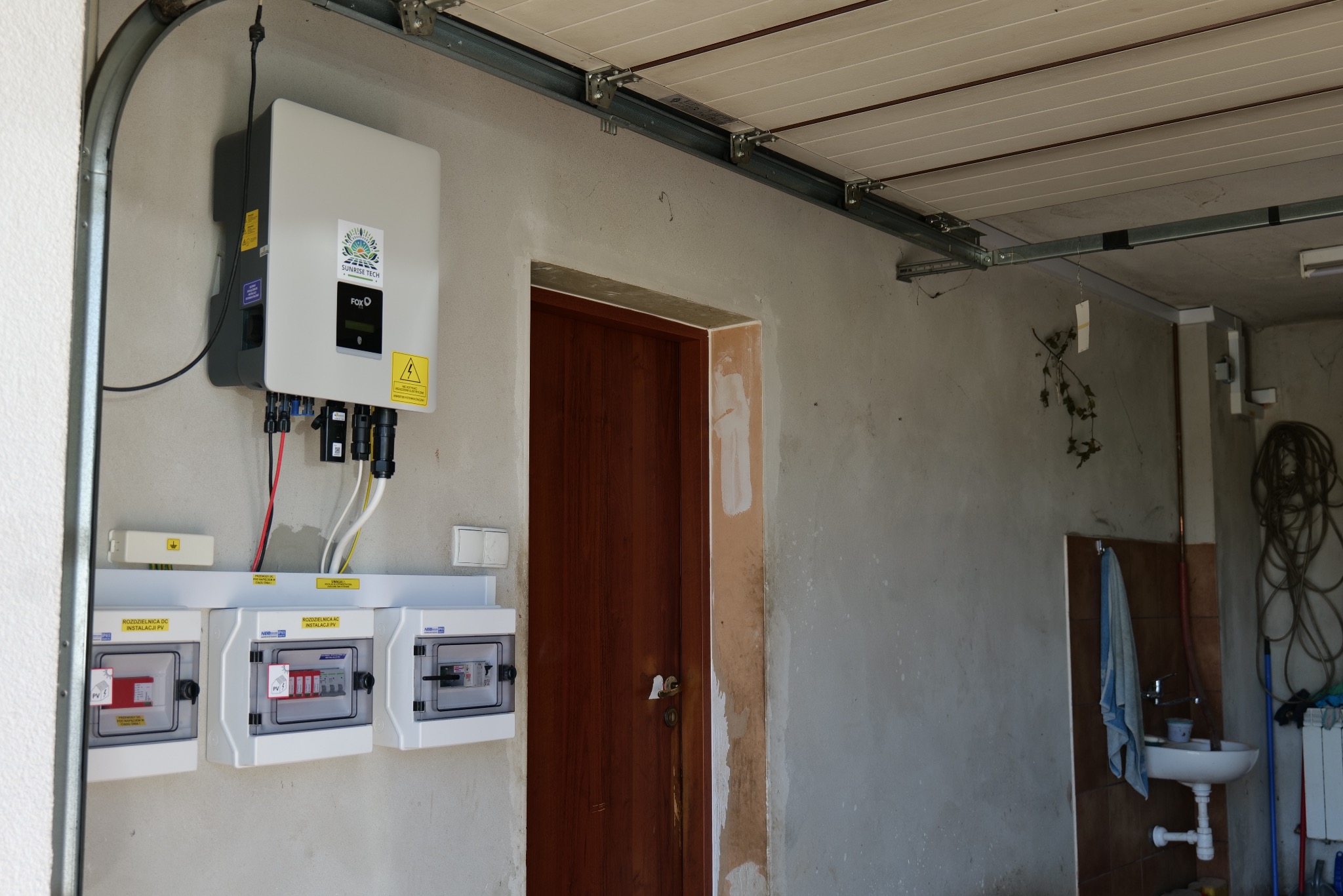 Instalacja fotowoltaiczna w garażu: falownik FOX ESS zawieszony na szarej ścianie, pod nim trzy skrzynki rozdzielcze DC i AC, widoczne przewody i elementy instalacji, w tle drzwi i umywalka.