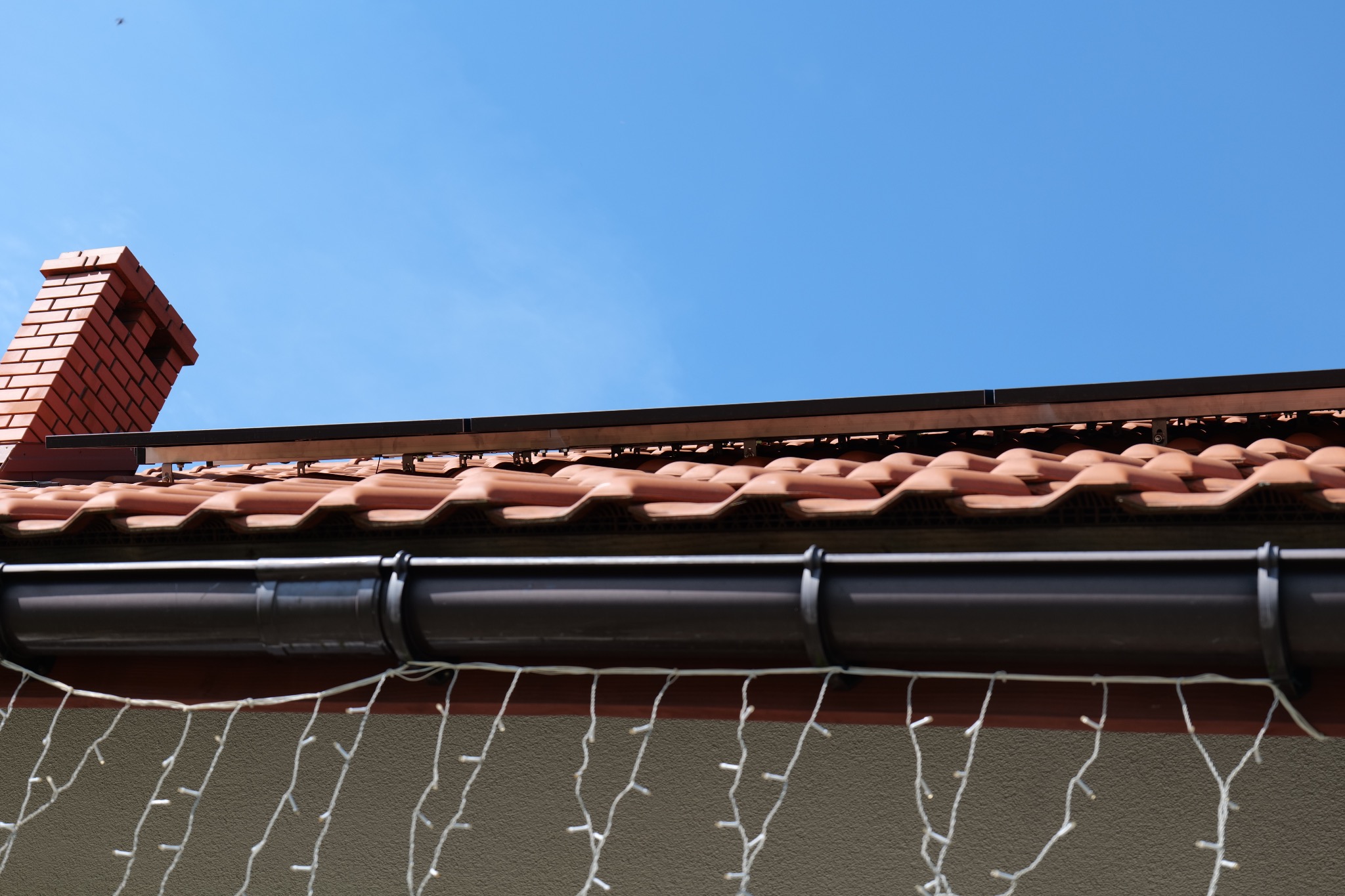 Czerwona dachówka ceramiczna z brązowymi rynnami i fragmentem instalacji pod panele, widoczny komin z cegły i girlanda świetlna.