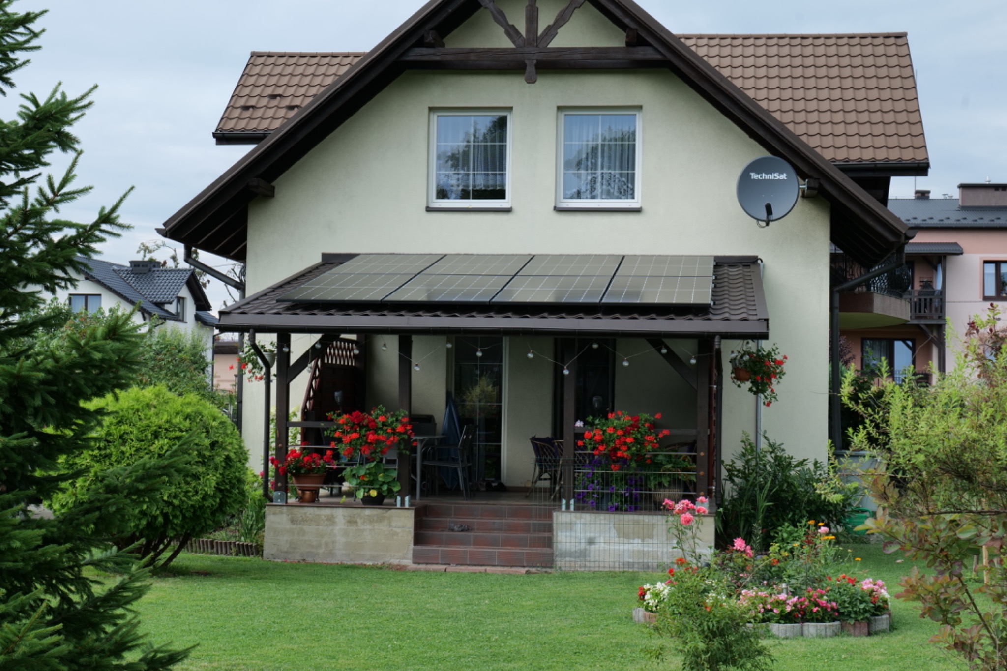 Dom jednorodzinny z panelami słonecznymi zamontowanymi nad werandą, widok z zadbanego ogrodu.