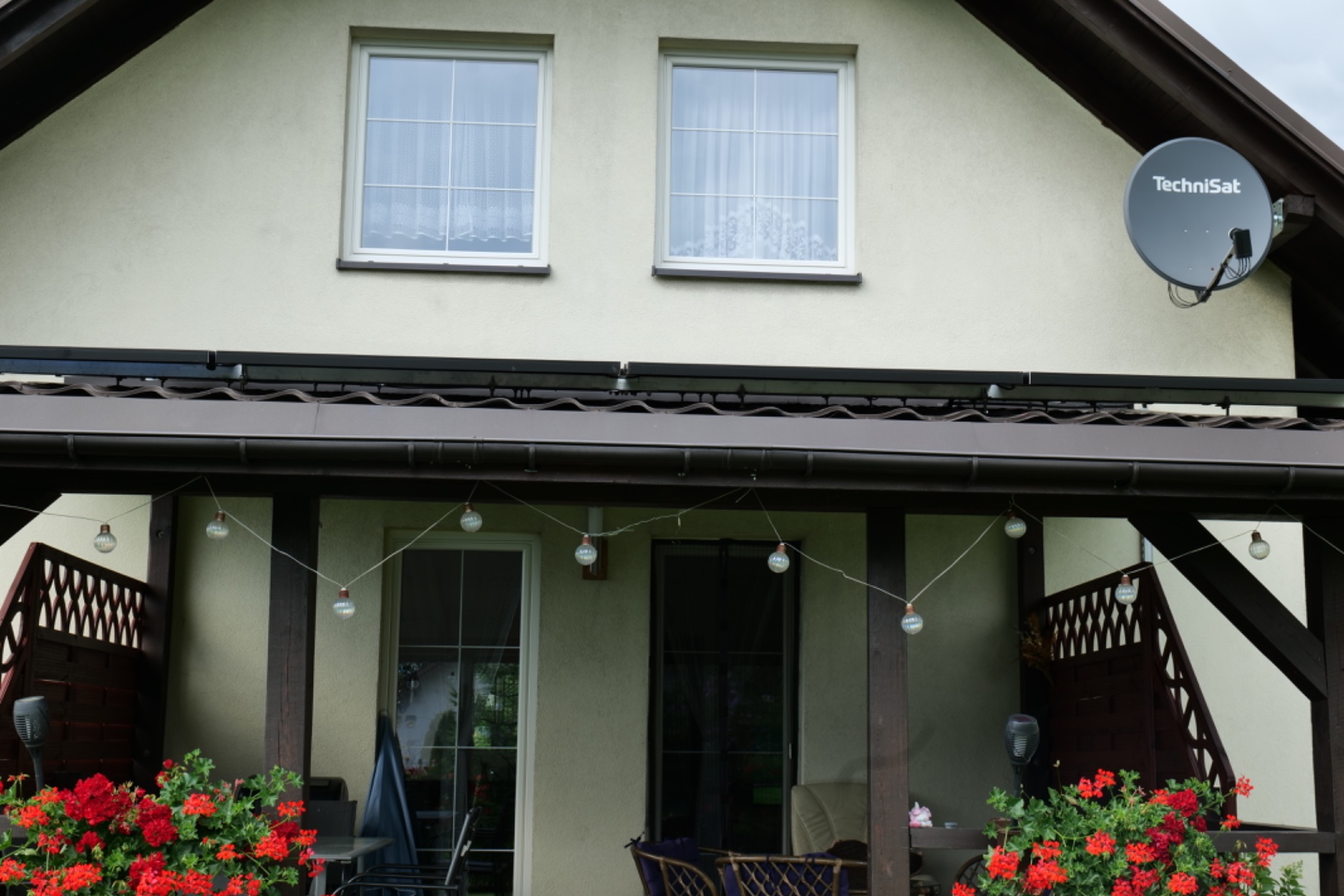 Dom jednorodzinny z panelami słonecznymi na dachu, widoczne zadaszenie tarasu z dekoracyjnymi żarówkami i kwiatami.