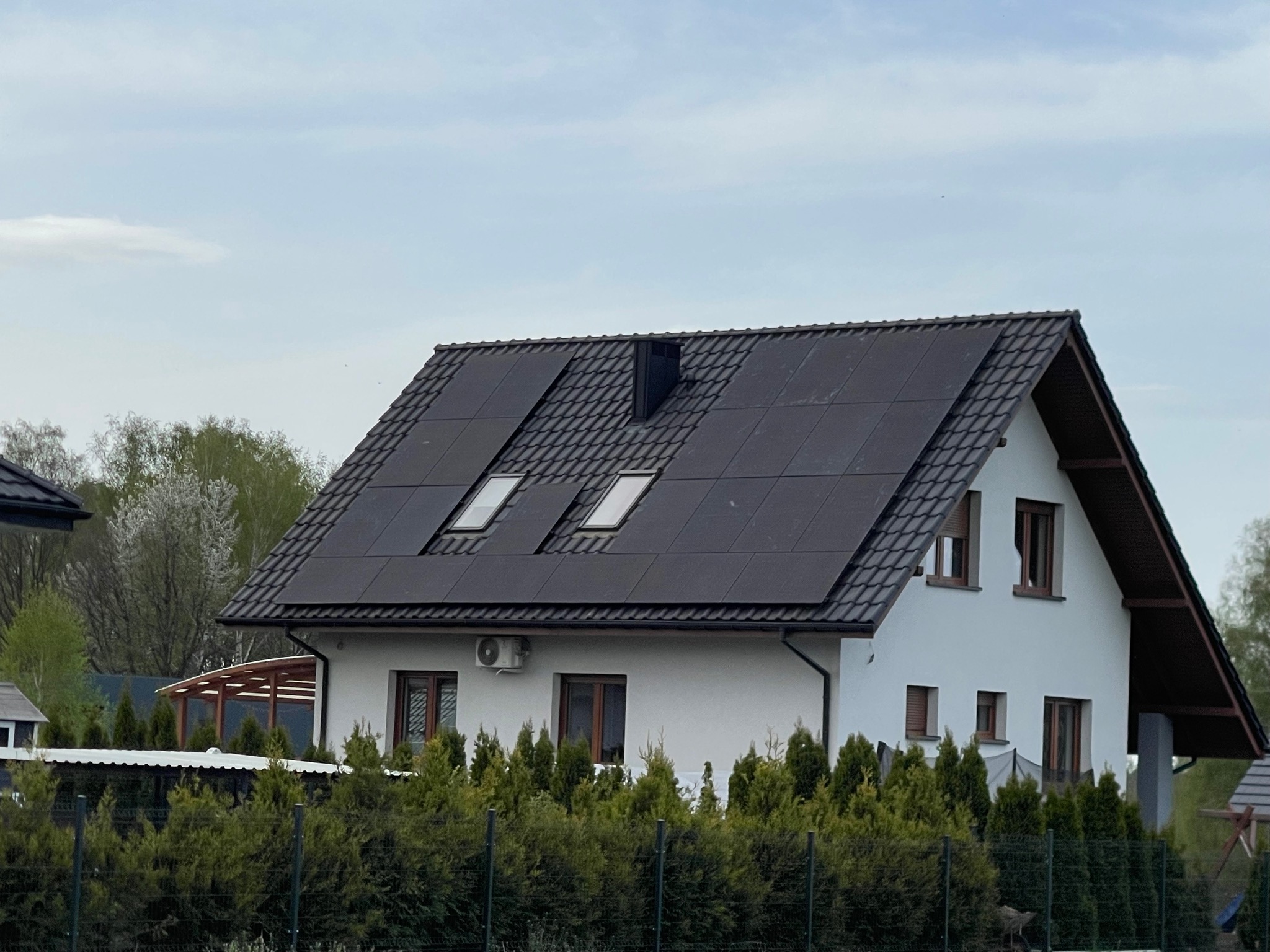 Biały dom z ciemną dachówką, na której zamontowano panele słoneczne i dwa okna dachowe. Dom otoczony zielenią, widok z poziomu ulicy.