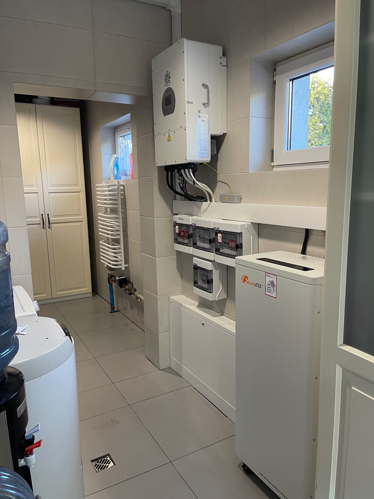 Biały inwerter fotowoltaiczny i magazyn energii Felicity ESS zamontowane w pomieszczeniu z białymi płytkami i szafkami. Widoczne okno i grzejnik.