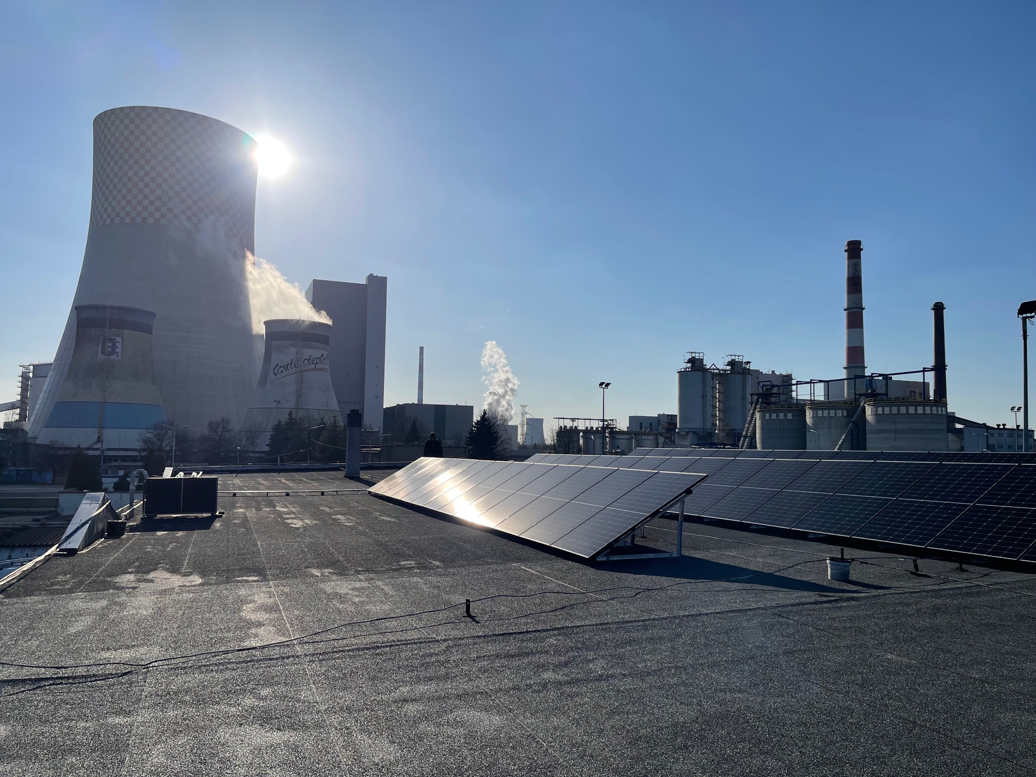 Instalacja paneli słonecznych na dachu budynku przemysłowego, w tle kominy i chłodnie kominowe. Słoneczny dzień.