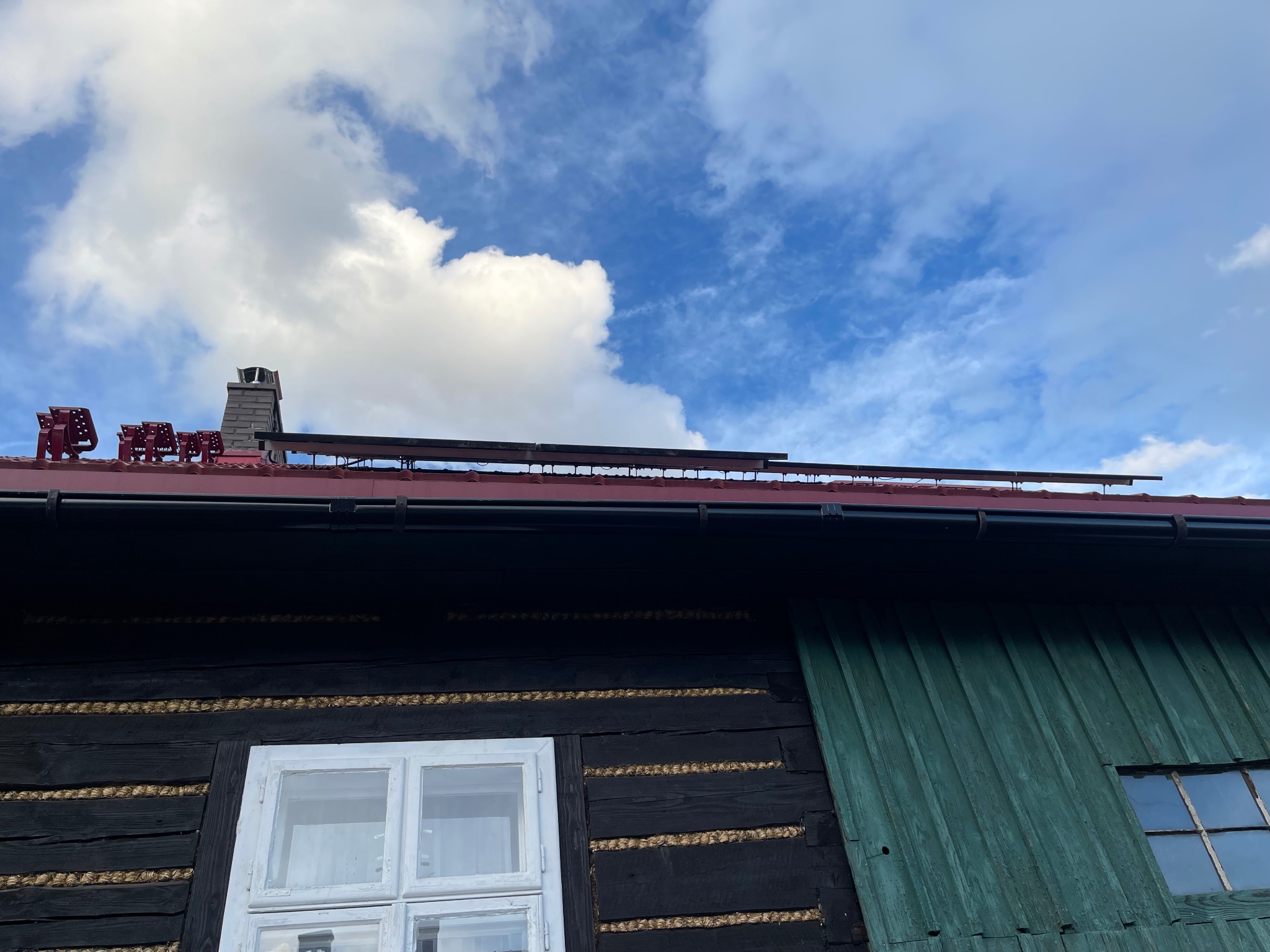 Panele fotowoltaiczne na dachu zabytkowego, drewnianego domu z oknami i kominem na tle błękitnego nieba z chmurami. Widok z dołu.