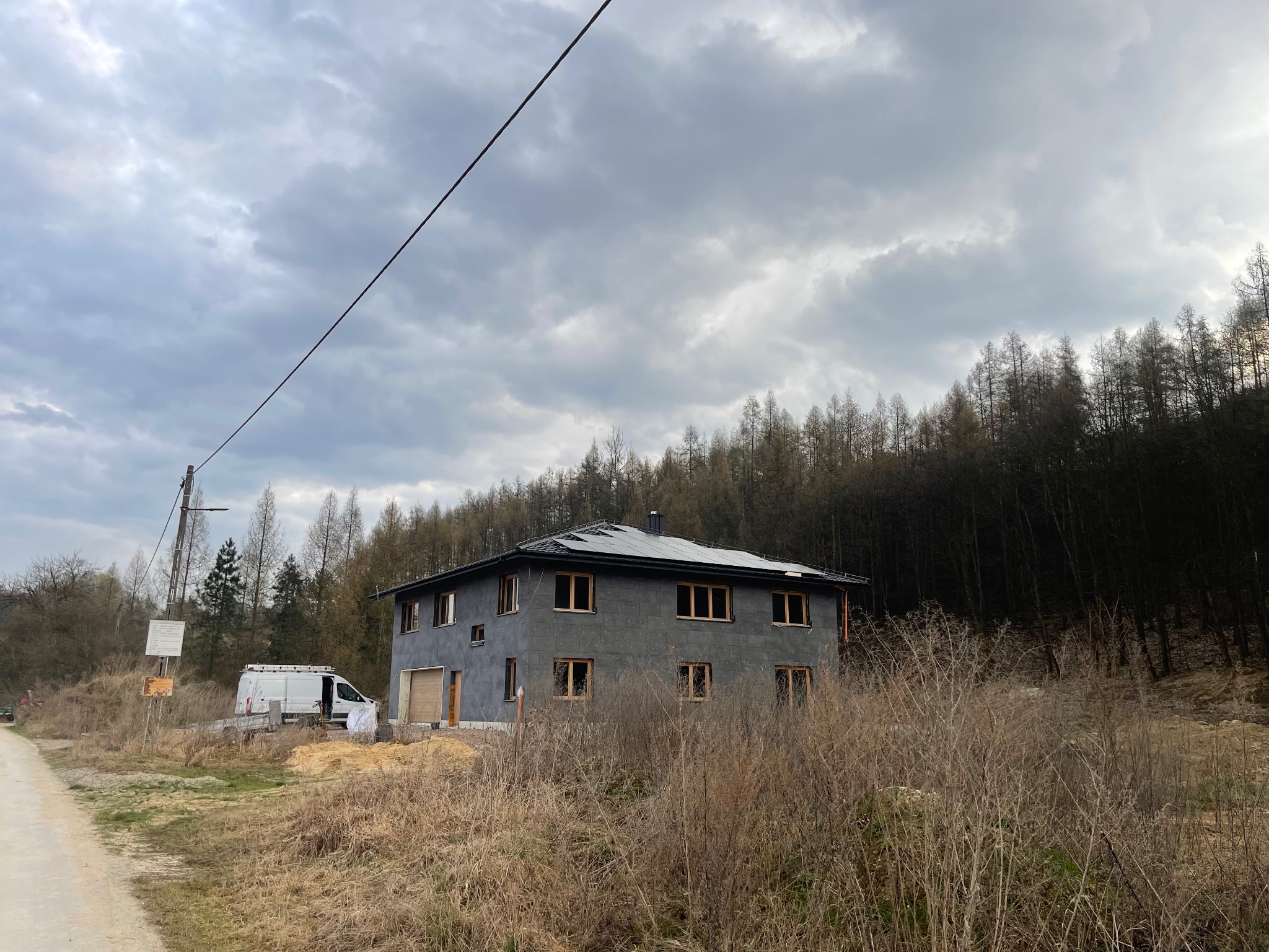 Szary dom z panelami słonecznymi na dachu w trakcie montażu, widoczny zarośnięty teren i samochód dostawczy w tle, pochmurne niebo.