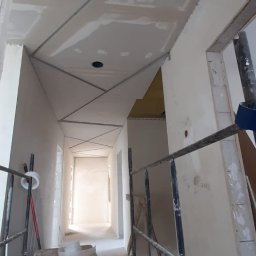 MDG - Korytarz w trakcie prac wykończeniowych: widoczny stelaż metalowy pod zabudowę sufitu z płyt, częściowo zamontowane płyty, otwór rewizyjny i drewniana drabinka strychowa, rusztowanie na pierwszym...