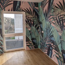 MDG - Pusty pokój z drewnianą podłogą i ścianą pokrytą tapetą w tropikalne liście na różowym tle, widok na okno.