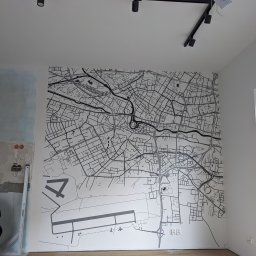 MDG - Ściana z mapą miasta z lotniskiem, czarno-biała grafika w nowym mieszkaniu z drewnianą podłogą, widoczne niedokończone prace na ścianie obok, czarne oświetlenie sufitowe.