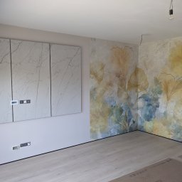 MDG - Pokój po malowaniu ścian z motywem liści ginkgo, z zamontowanymi szafkami z marmurowym wzorem i odkrytymi gniazdkami elektrycznymi, podłoga w jasnym odcieniu drewna.