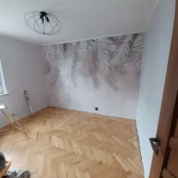 Pokój po odświeżeniu: parkiet w jodełkę, ściana z motywem liści palmowych, nowoczesna lampa sufitowa, drabina oparta o ścianę.