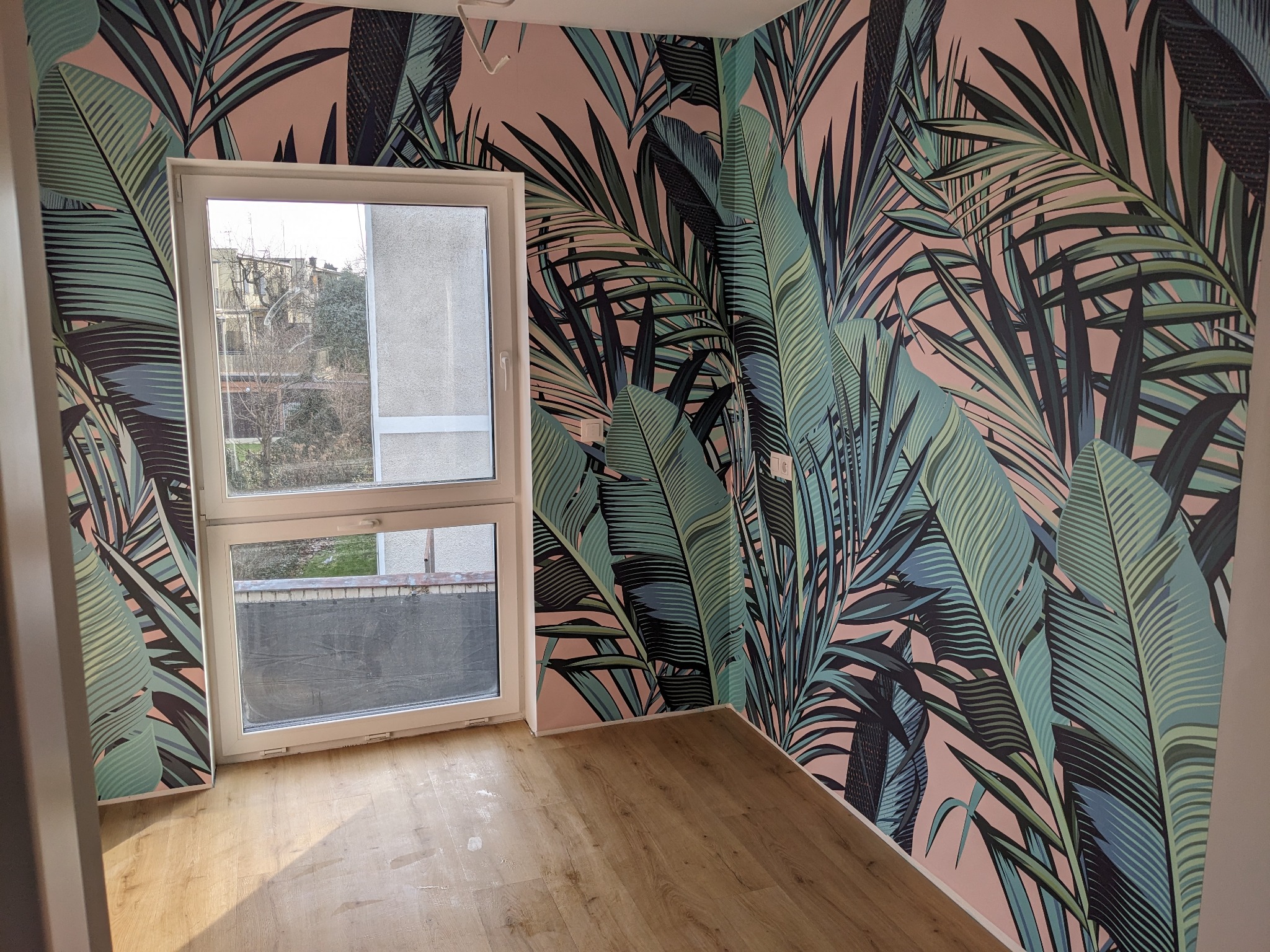 Pusty pokój z drewnianą podłogą i ścianą pokrytą tapetą w tropikalne liście na różowym tle, widok na okno.