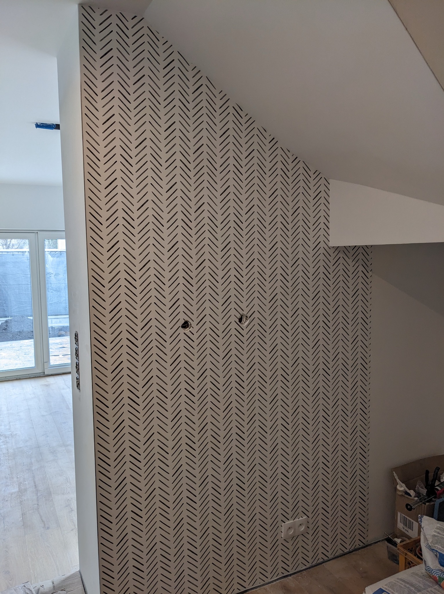 Ściana wykończona płytą kartonowo-gipsową z dekoracyjnym wzorem w jodełkę, widoczne otwory na gniazdka elektryczne, fragment podłogi z panelami i okno balkonowe w tle.