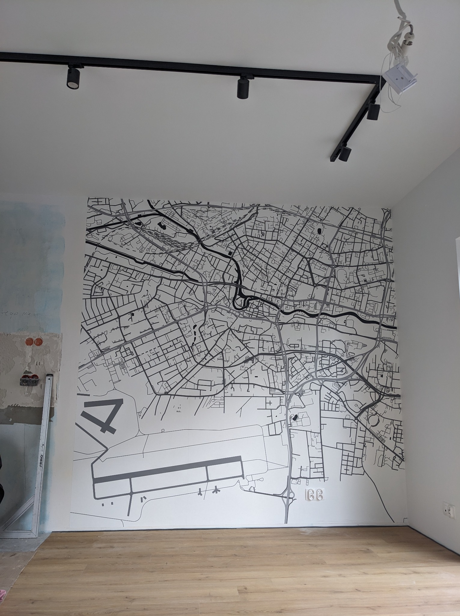 Ściana z mapą miasta z lotniskiem, czarno-biała grafika w nowym mieszkaniu z drewnianą podłogą, widoczne niedokończone prace na ścianie obok, czarne oświetlenie sufitowe.