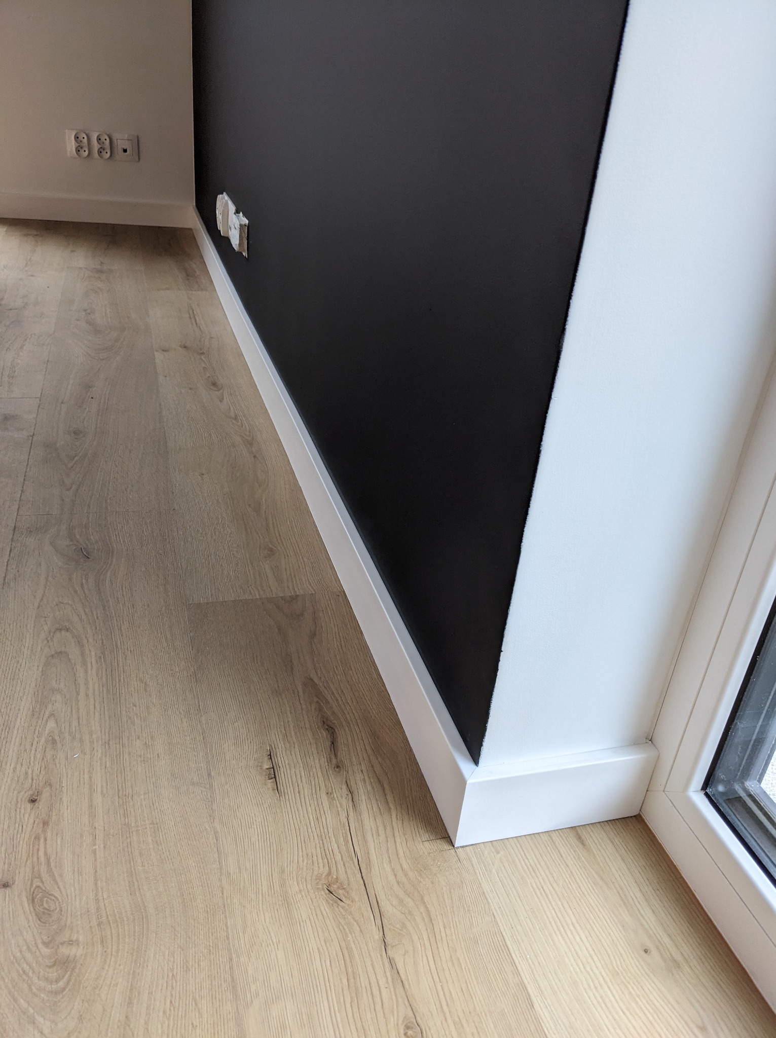 Fragment wykończonego wnętrza w Bydgoszczy: ciemna ściana kontrastuje z jasną podłogą laminowaną i białymi listwami przypodłogowymi, widoczne gniazdka elektryczne.