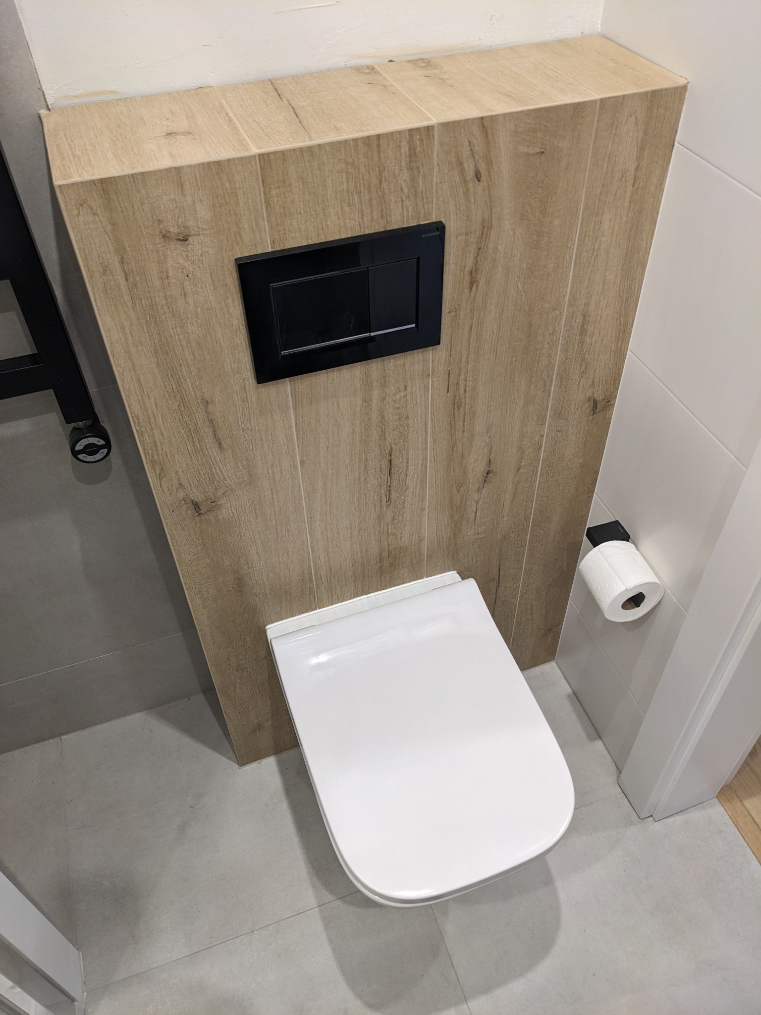 Wisząca toaleta z czarnym panelem spłukującym, obudowana płytkami imitującymi drewno, z widocznym uchwytem na papier toaletowy i szarą podłogą.