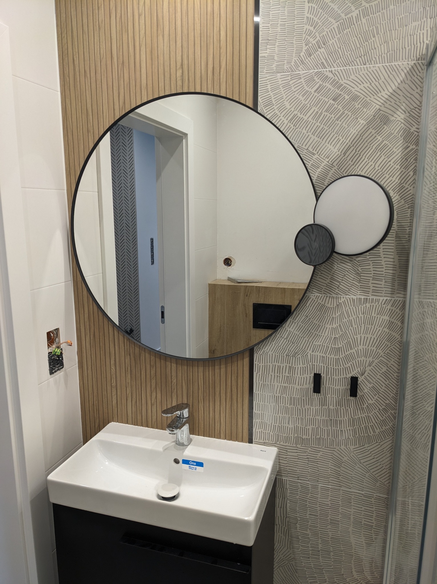 Wykończona łazienka z okrągłym lustrem z podświetleniem, białą umywalką Roca na czarnej szafce, drewnopodobną i wzorzystą szarą okładziną ścienną oraz widocznym fragmentem kabiny prysznicowej.