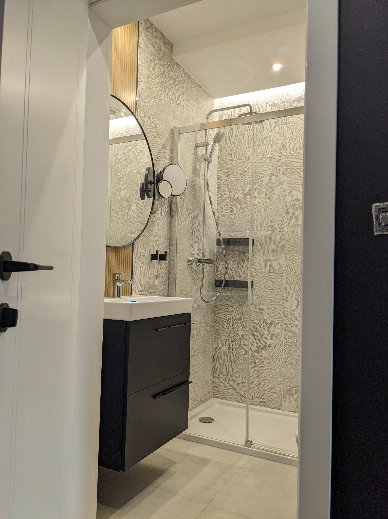 Nowoczesna łazienka z kabiną prysznicową, umywalką z szafką w kolorze grafitowym, okrągłym lustrem i płytkami imitującymi rysunek drewna na ścianach, widziana z perspektywy otwartych drzwi.