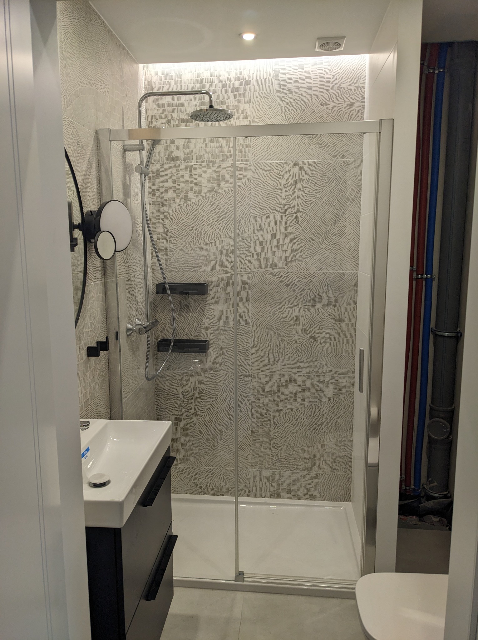 Nowoczesna kabina prysznicowa z deszczownicą, umywalką z szafką w wykończonej łazience, widoczne rury instalacji wodnej.